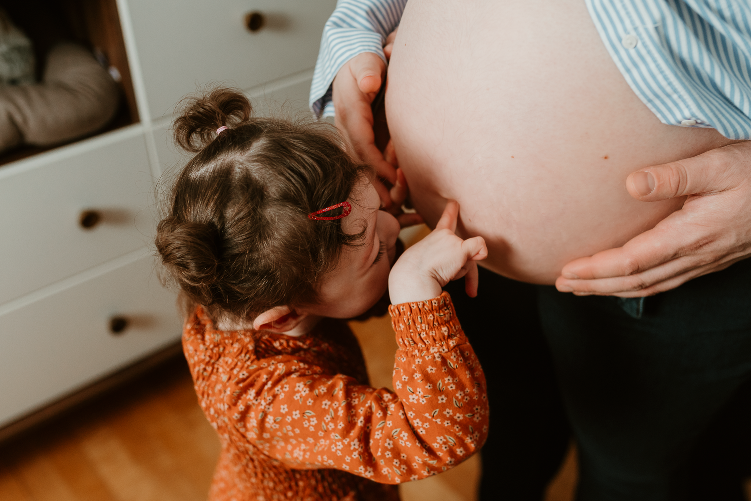 Young girl in orange dress kissing pregnant mother's baby bump in a warm, heartfelt maternity photo.