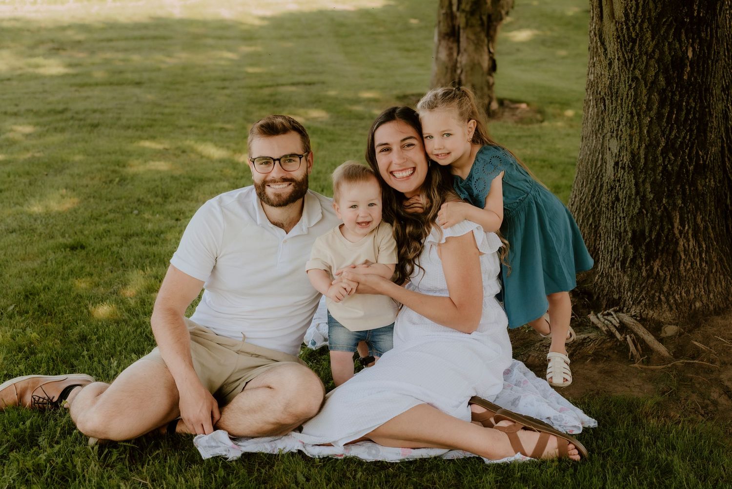A family group sits together on a grassy lawn under a large tree during an outdoor photo session.