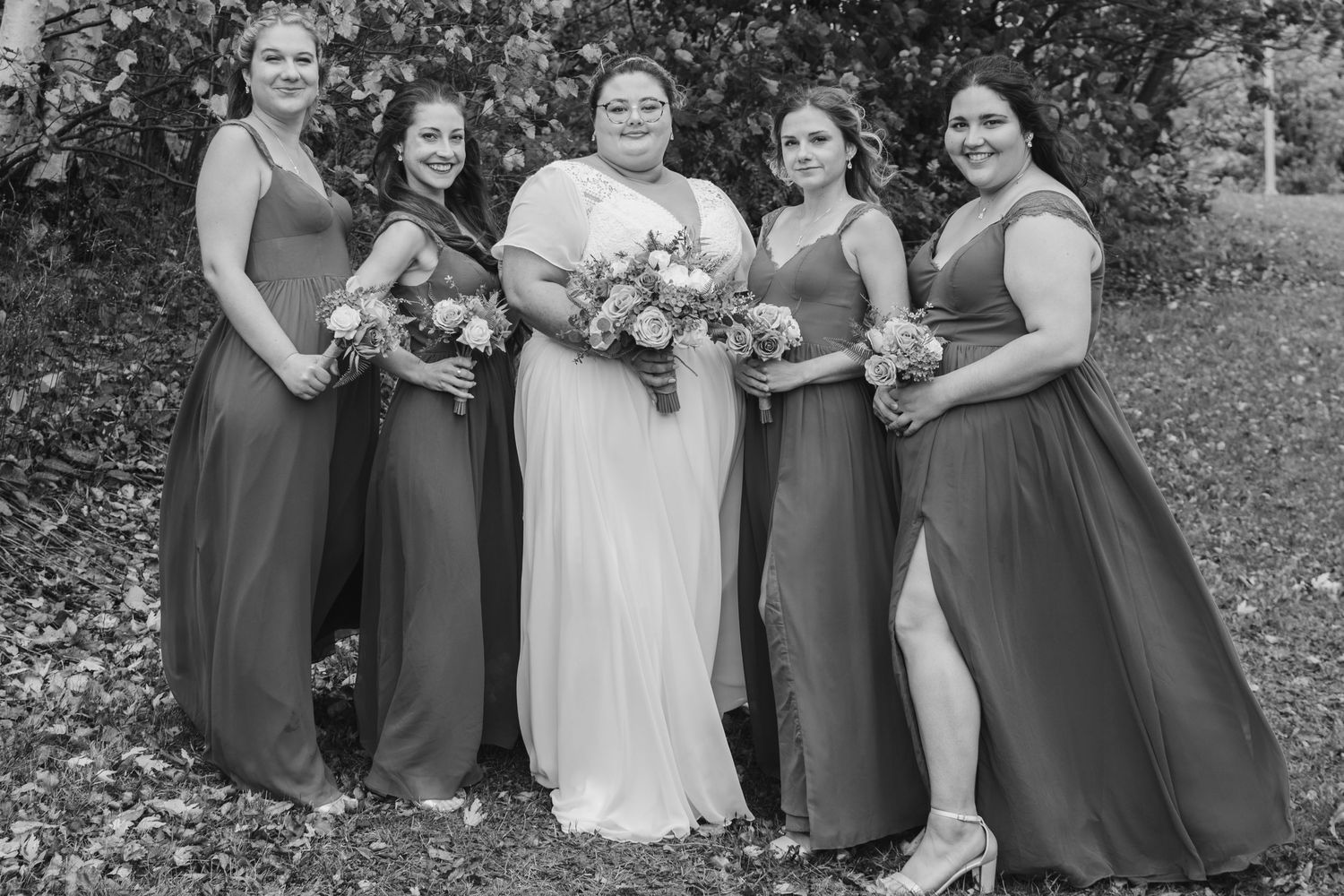 Bridal party in matching burgundy dresses poses outdoors for wedding photo.