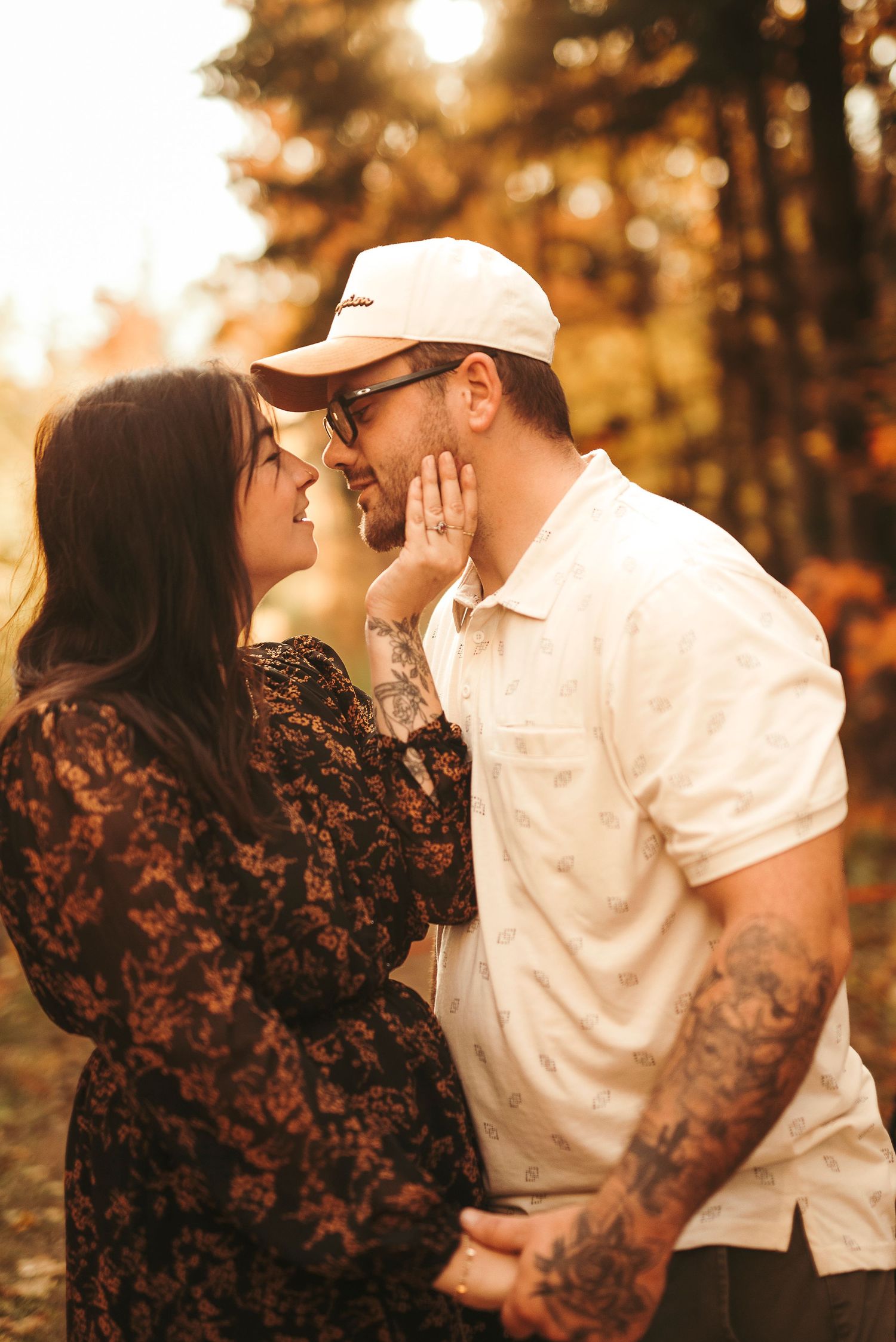 Couple sharing a tender moment outdoors in autumn, woman in lace dress touching mans face, both smiling, fall foliage background.