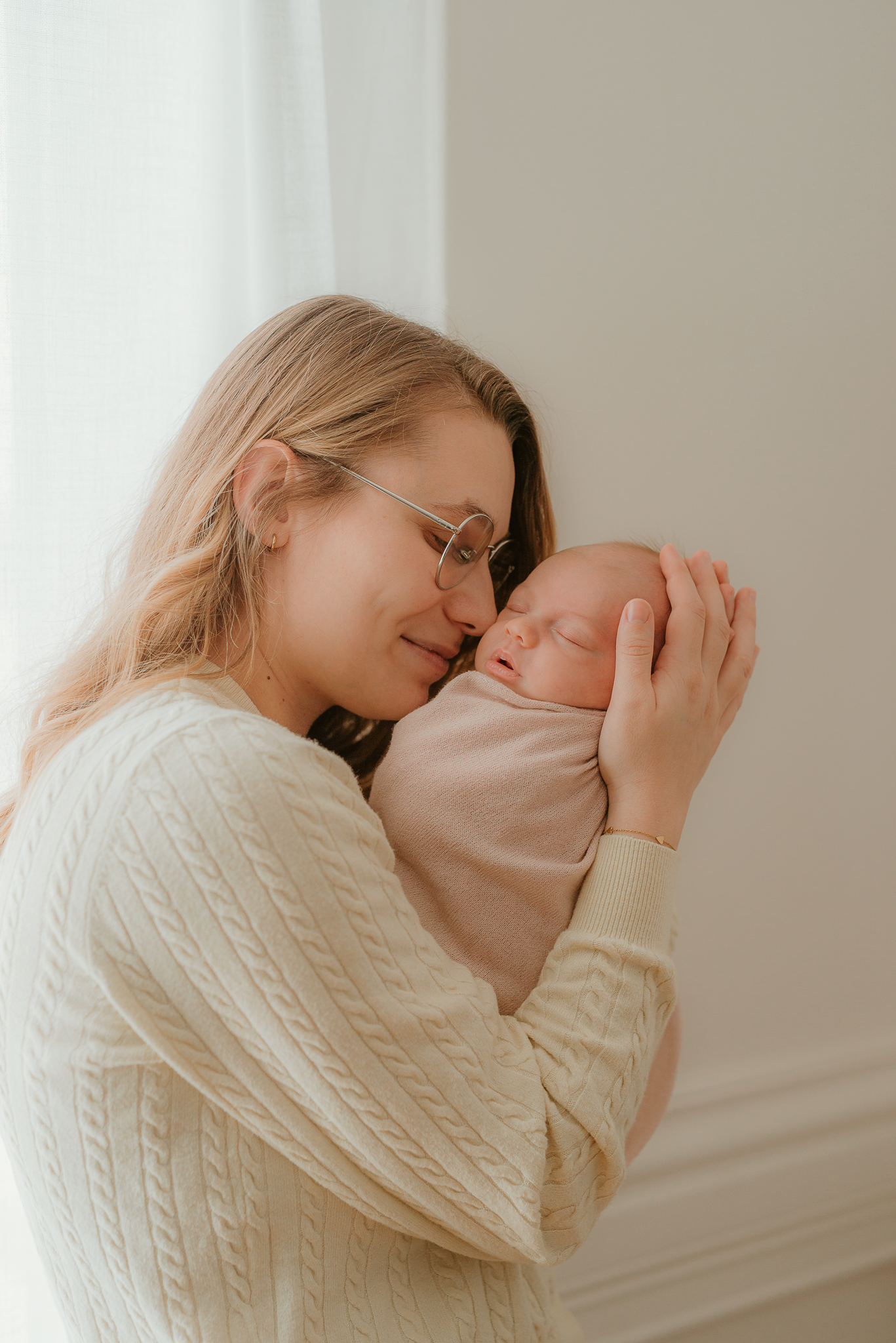 A mother in a cream cable-knit sweater tenderly holds her sleeping newborn baby, touching foreheads in a bright room.