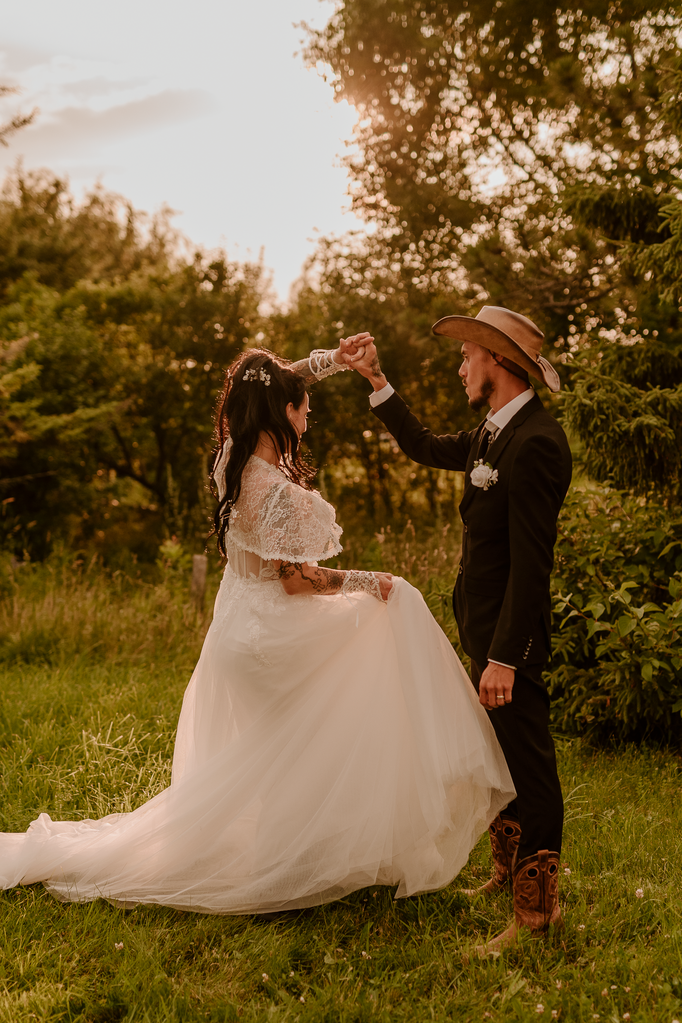 A bride and groom dance outdoors at golden hour, she in a lace gown, he in a suit and cowboy hat.
