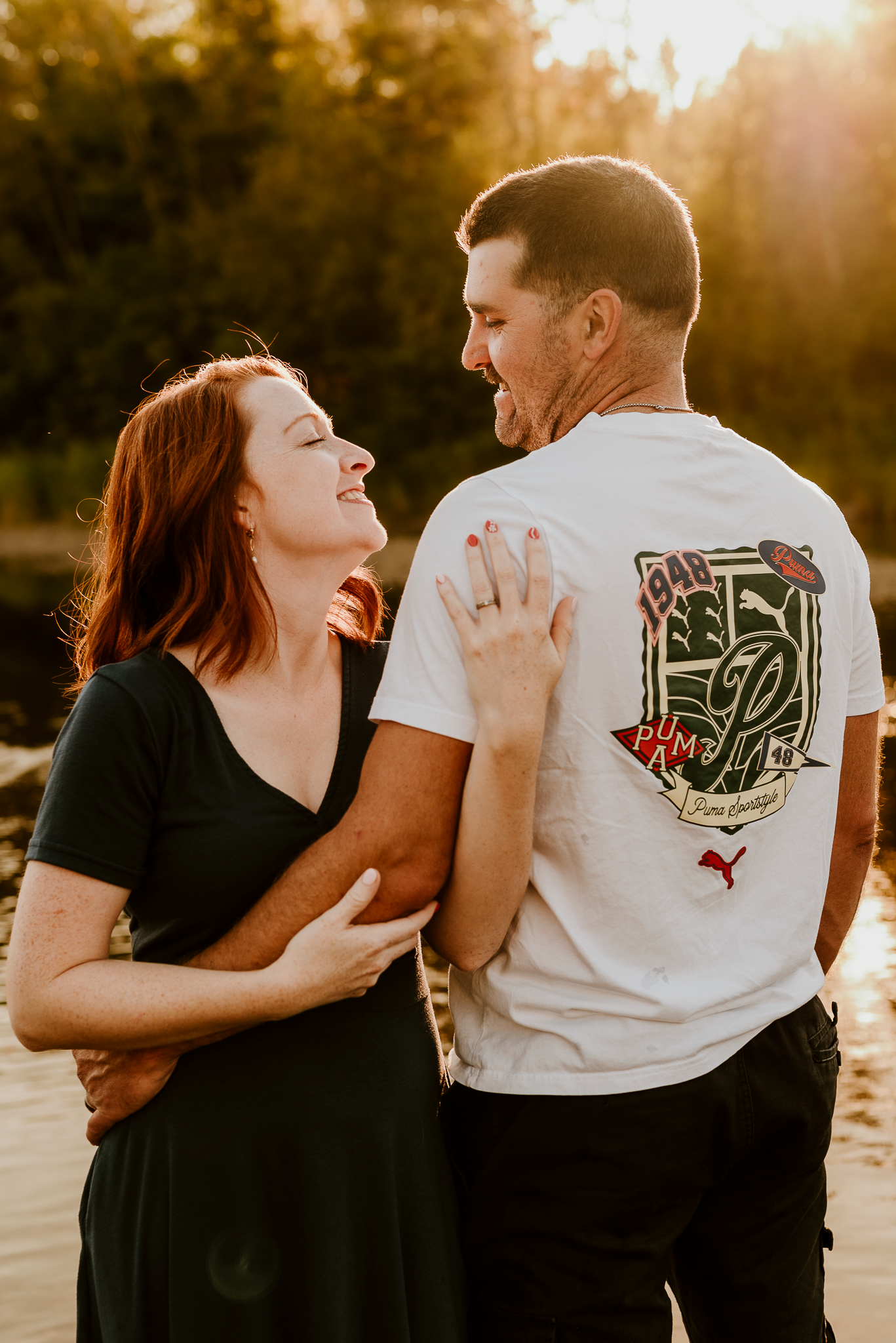 A couple laughs together at golden hour, woman in black dress, man in white Puma tee, outdoors near water.