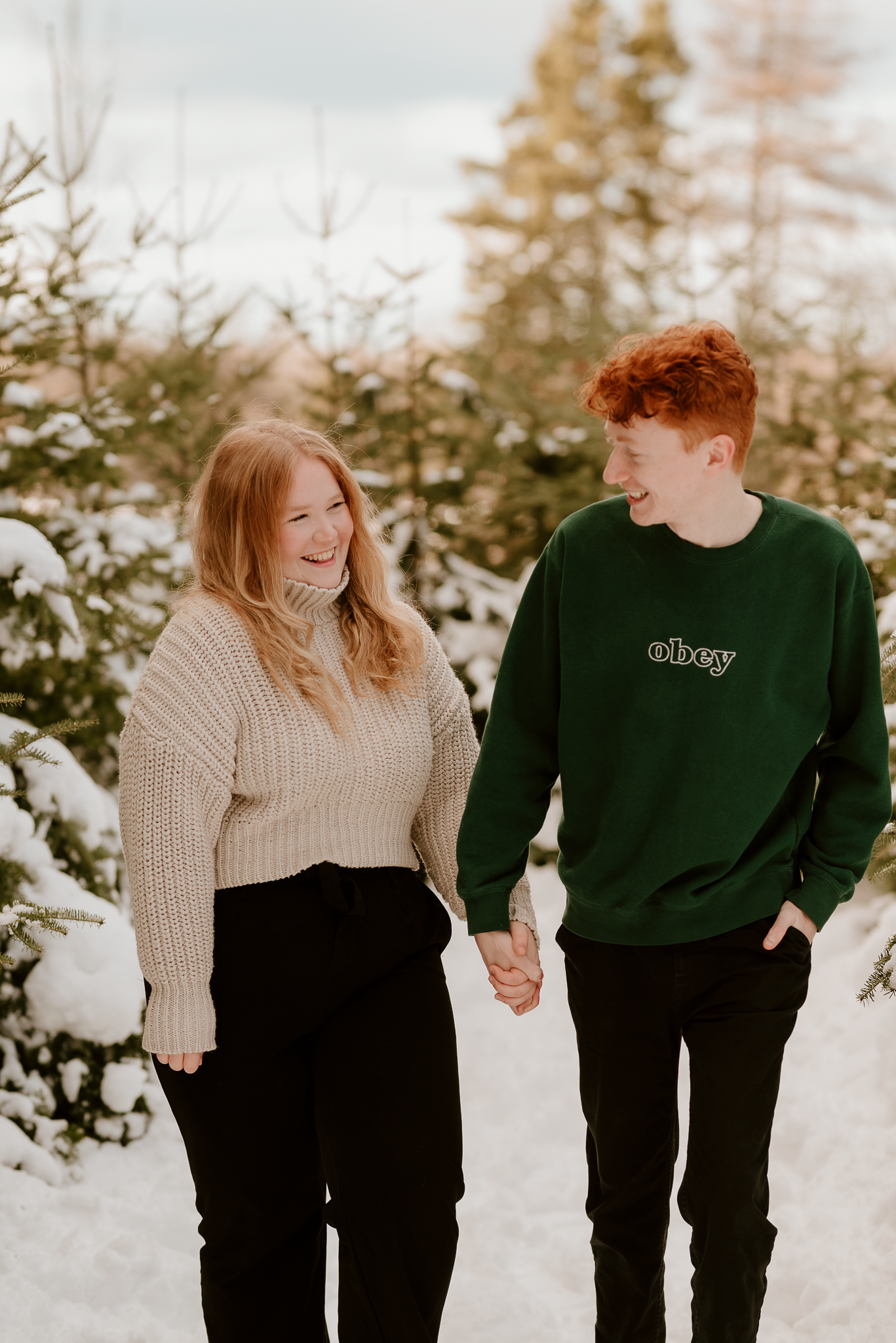A smiling couple holding hands in a snowy Christmas tree farm, wearing cozy sweaters in winter.