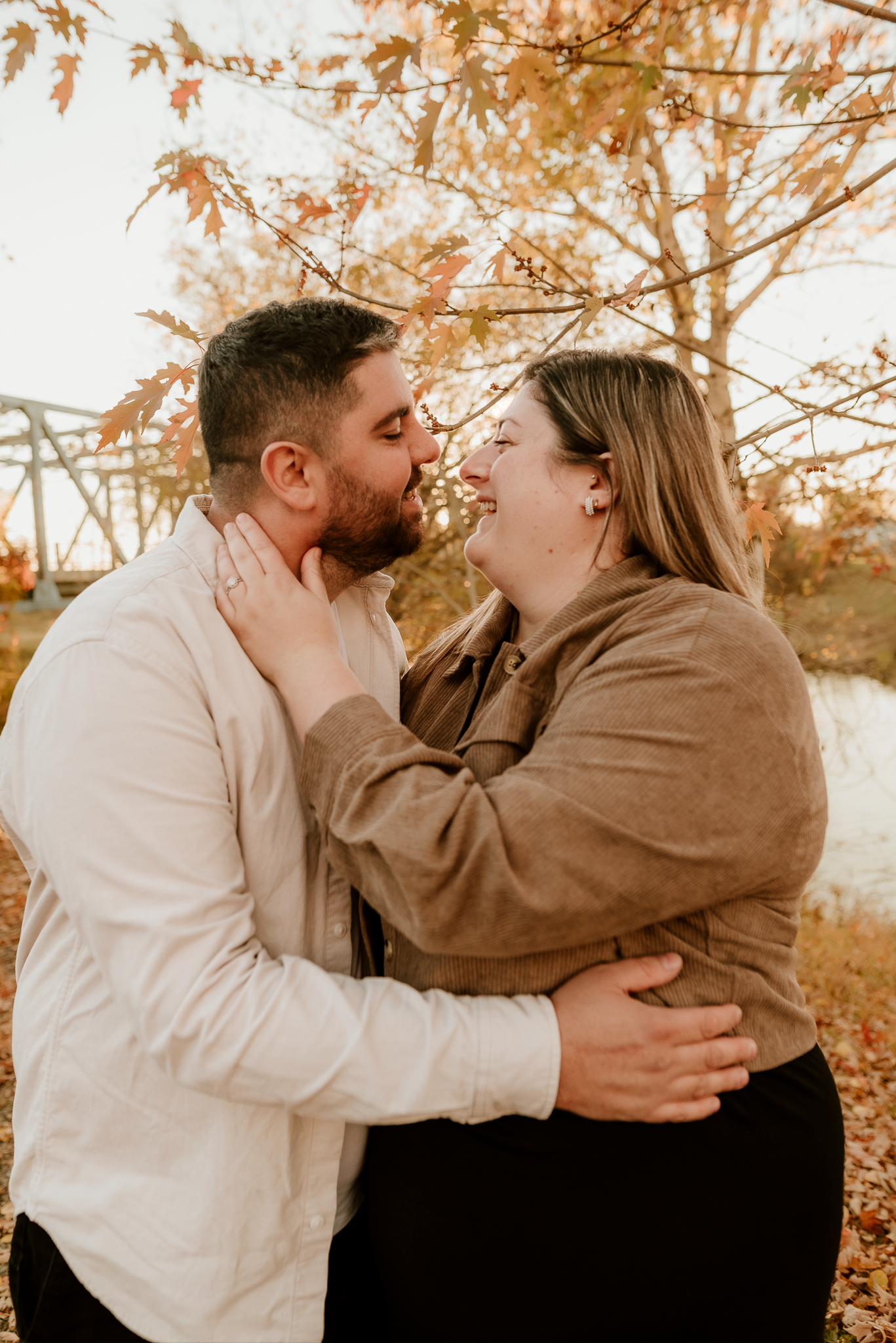 Couple sharing a romantic kiss outdoors in autumn, surrounded by golden fall foliage near a bridge.