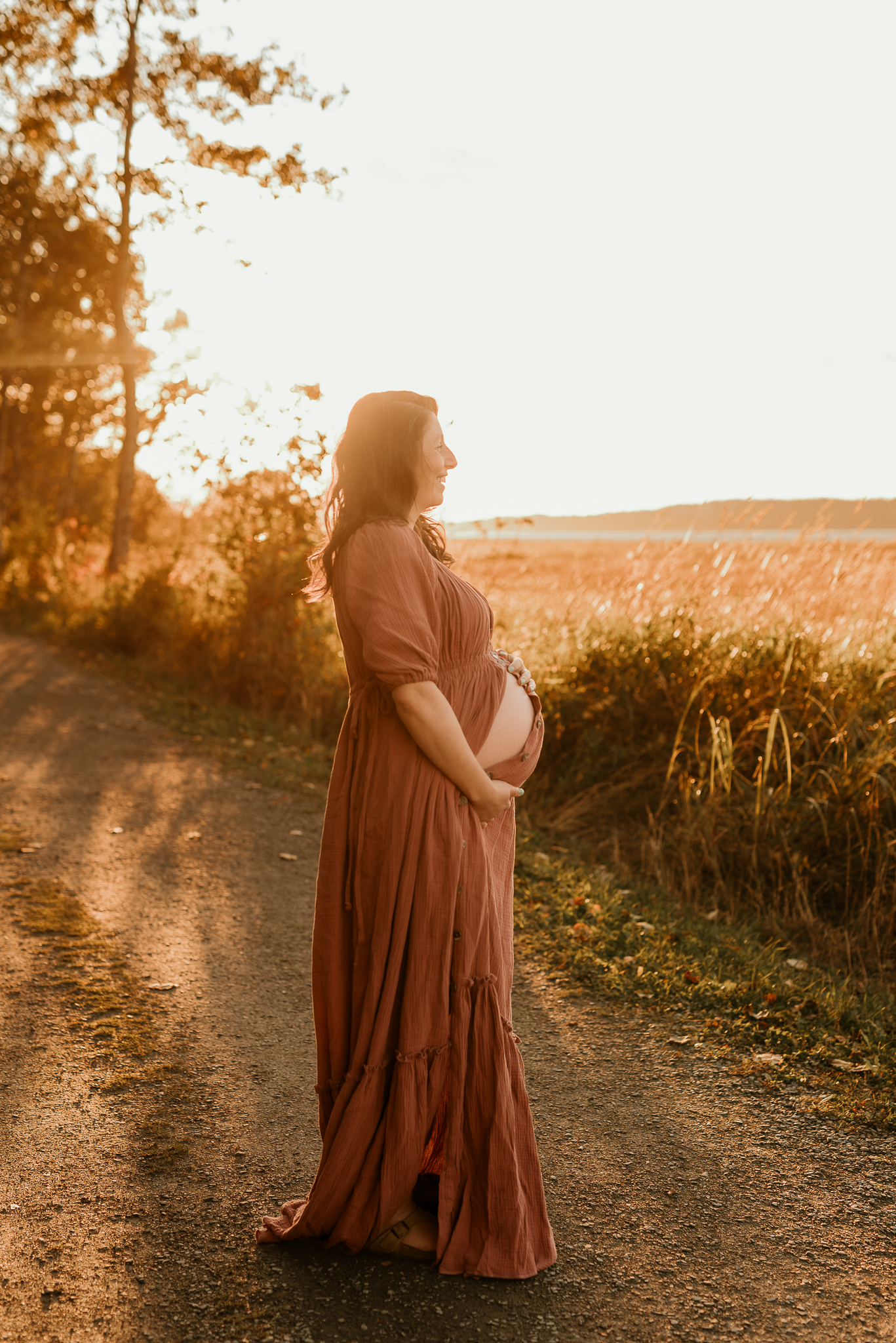 Pregnant woman in rust maxi dress holding baby bump on a sunlit country road during golden hour.