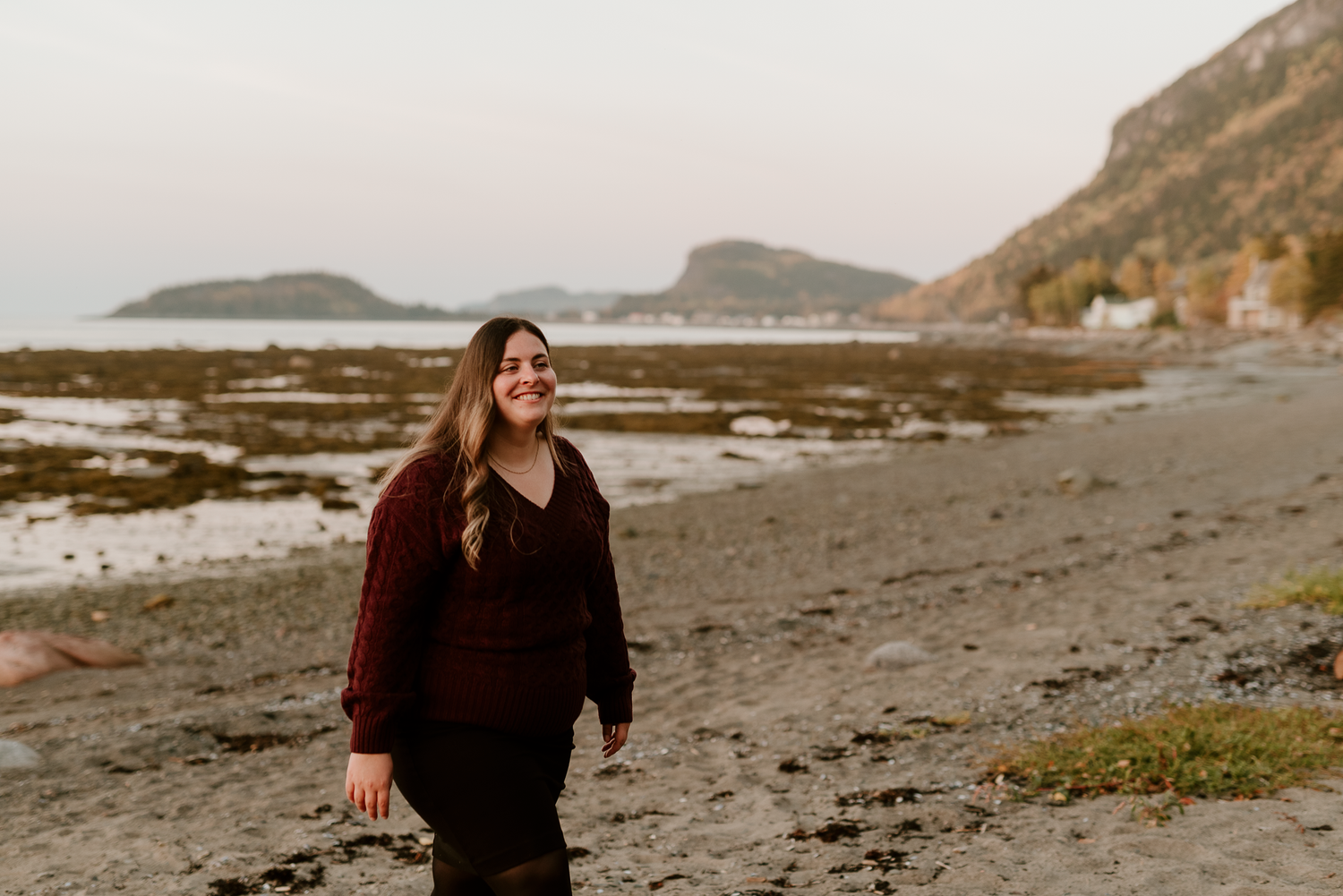 A smiling woman in a burgundy sweater walks along a rocky beach with misty mountains in the background.