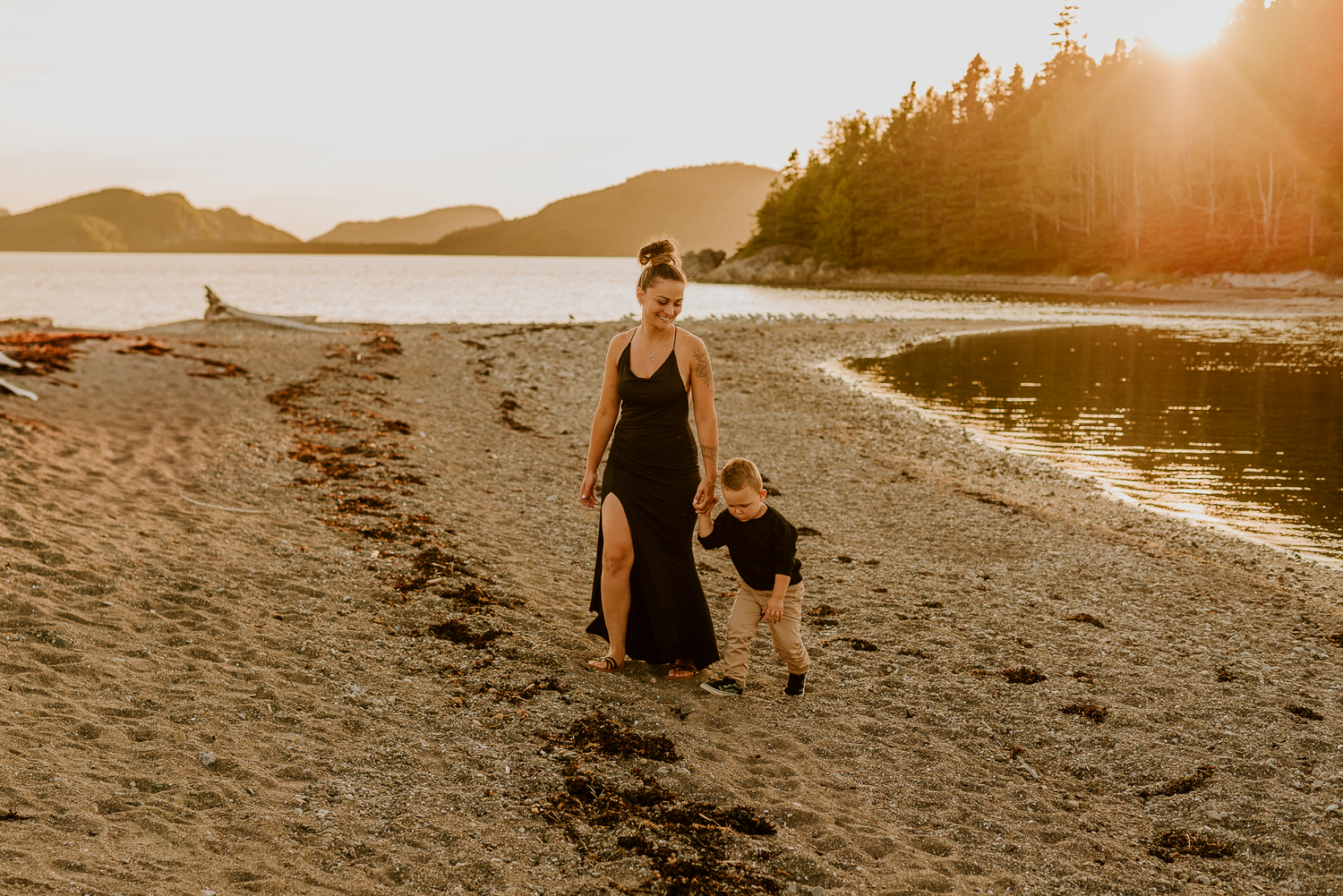 Mother and toddler walking hand in hand on a sandy beach at golden sunset with mountains and trees in background.