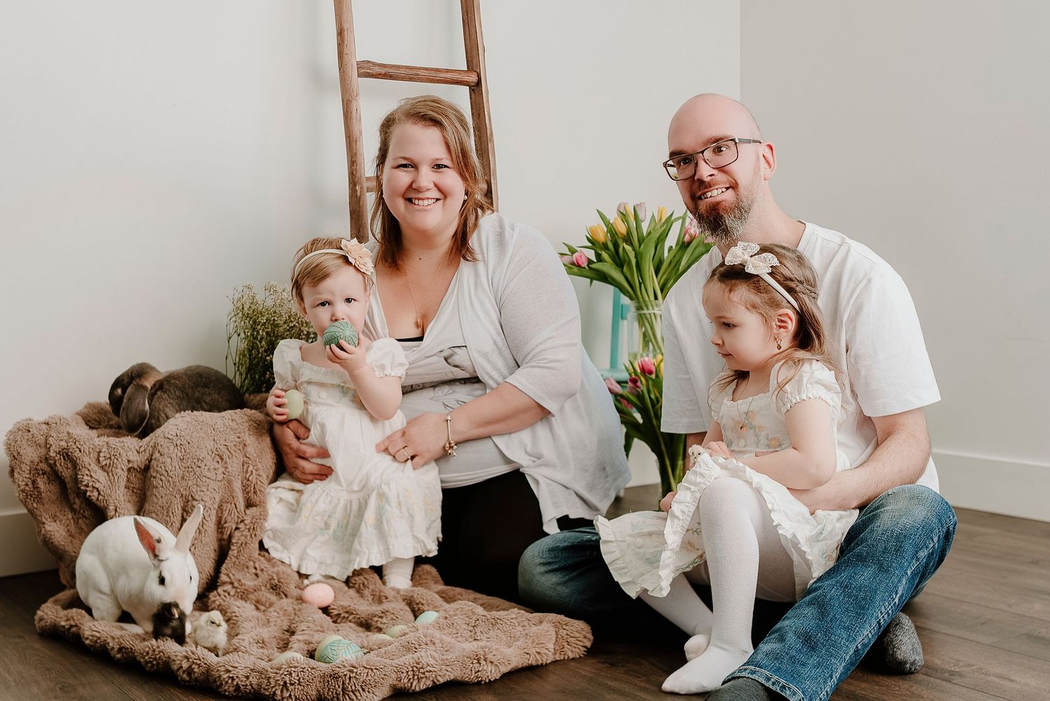 Family of four posing for Easter photos with white bunny, tulips, and rustic ladder backdrop in bright studio.