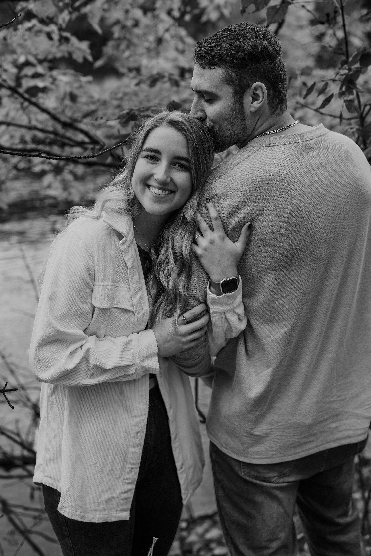 Une photo en noir et blanc d'une jeune femme enlaçant son amoureux, qui lui dépose un baiser sur sa tête, le tout dans un décor extérieur.