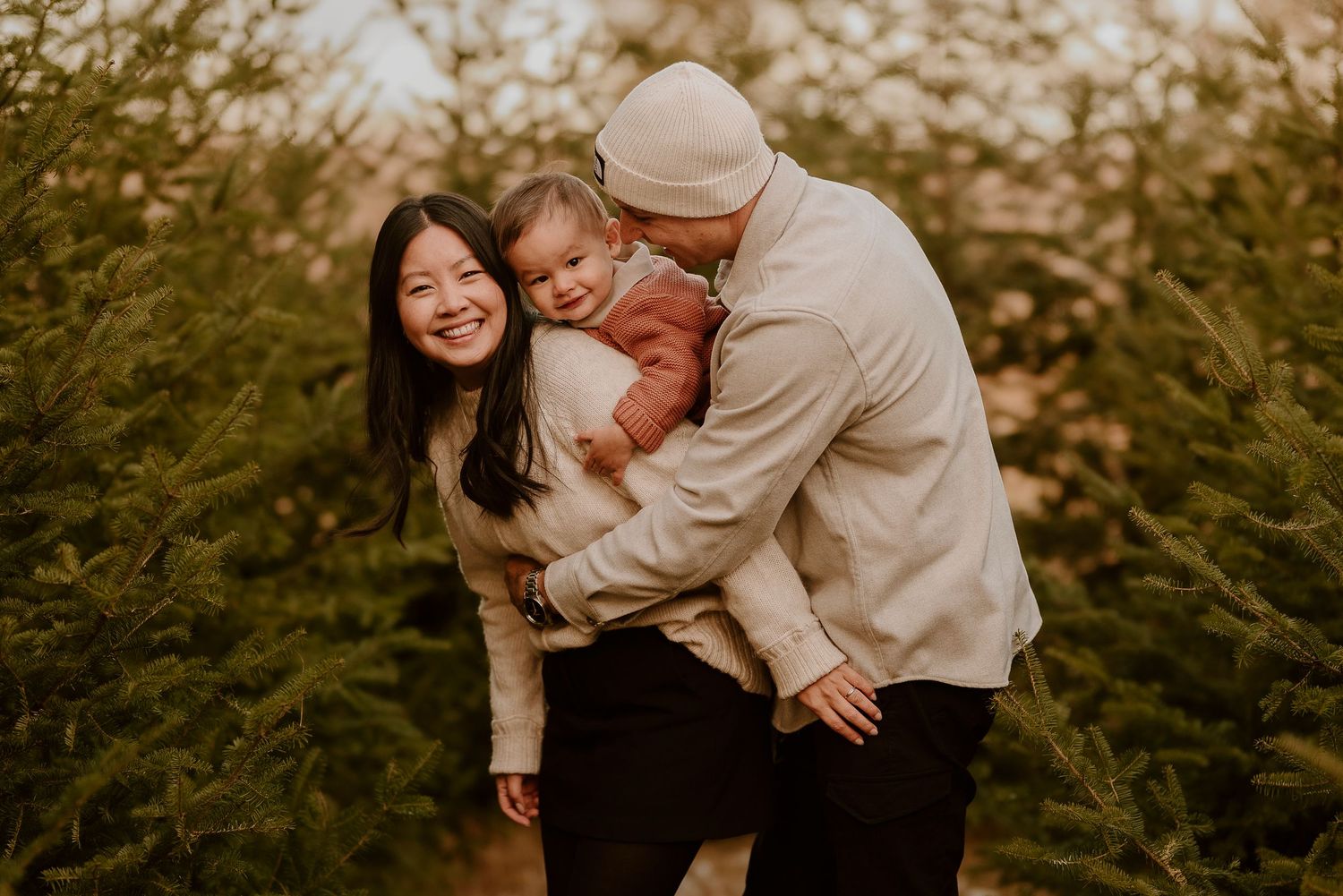 A happy family poses among evergreen trees, with a smiling mother, father in beanie, and baby in a warm outdoor portrait.