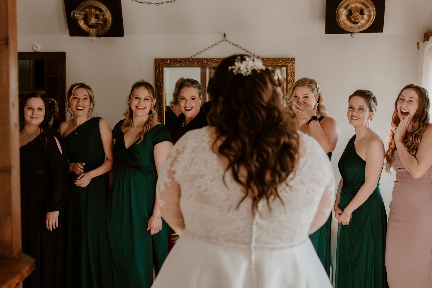 Bride reveals wedding dress to bridesmaids in green and pink gowns, capturing joyful reactions during bridal party getting ready.