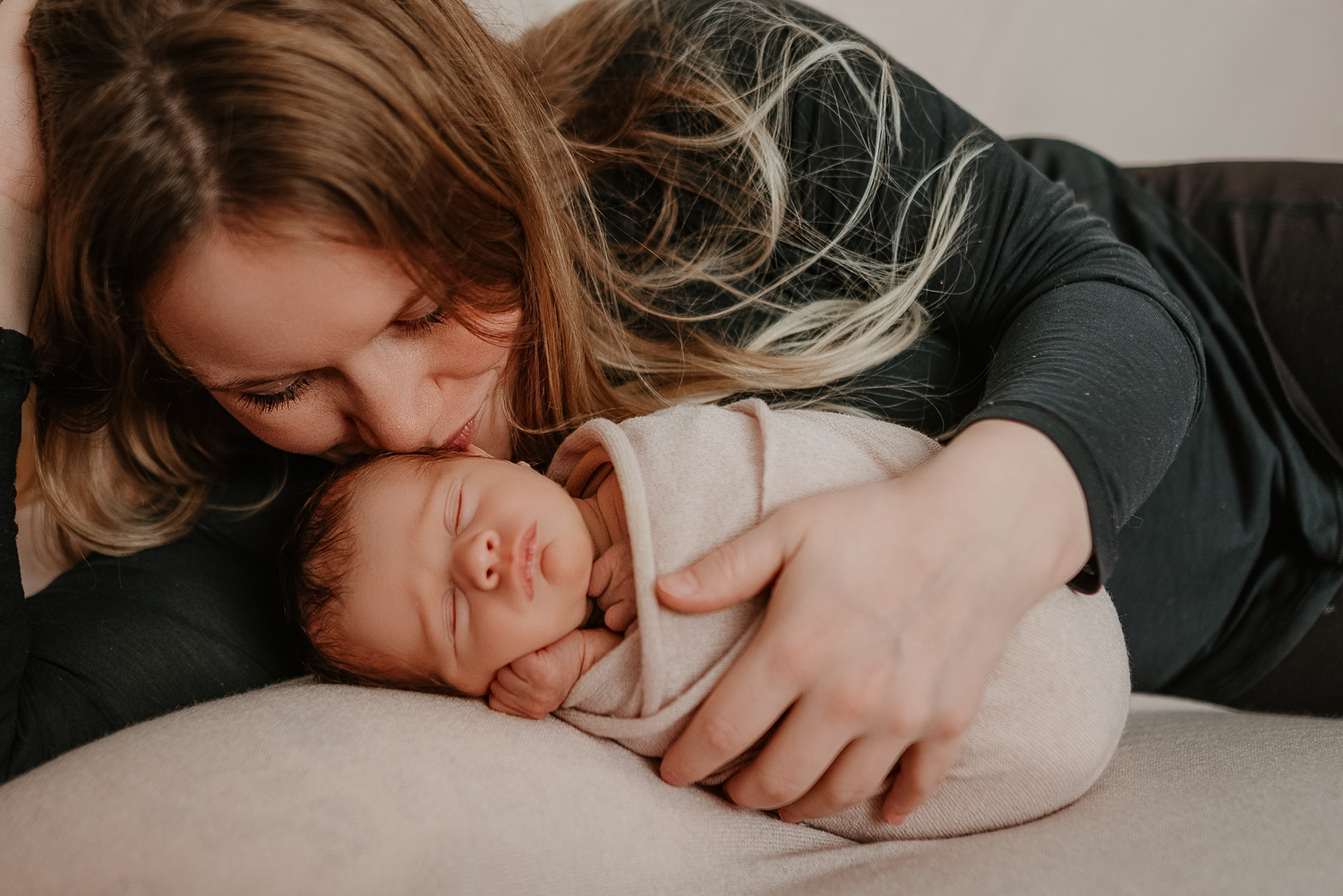 A mother tenderly kisses her sleeping swaddled newborn baby lying on a soft surface.