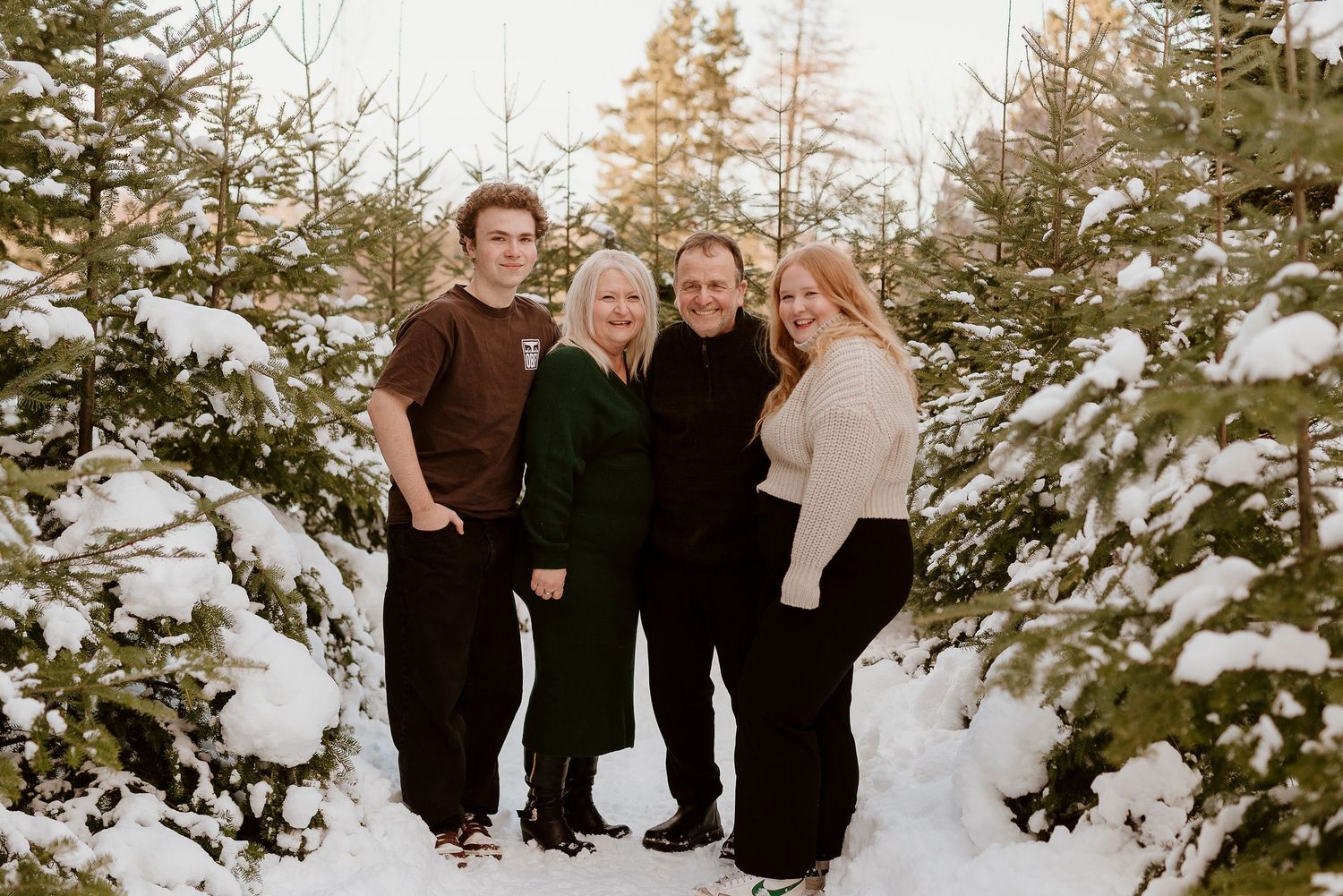 Family of four smiling together in a snowy Christmas tree farm, surrounded by snow-covered evergreen trees.