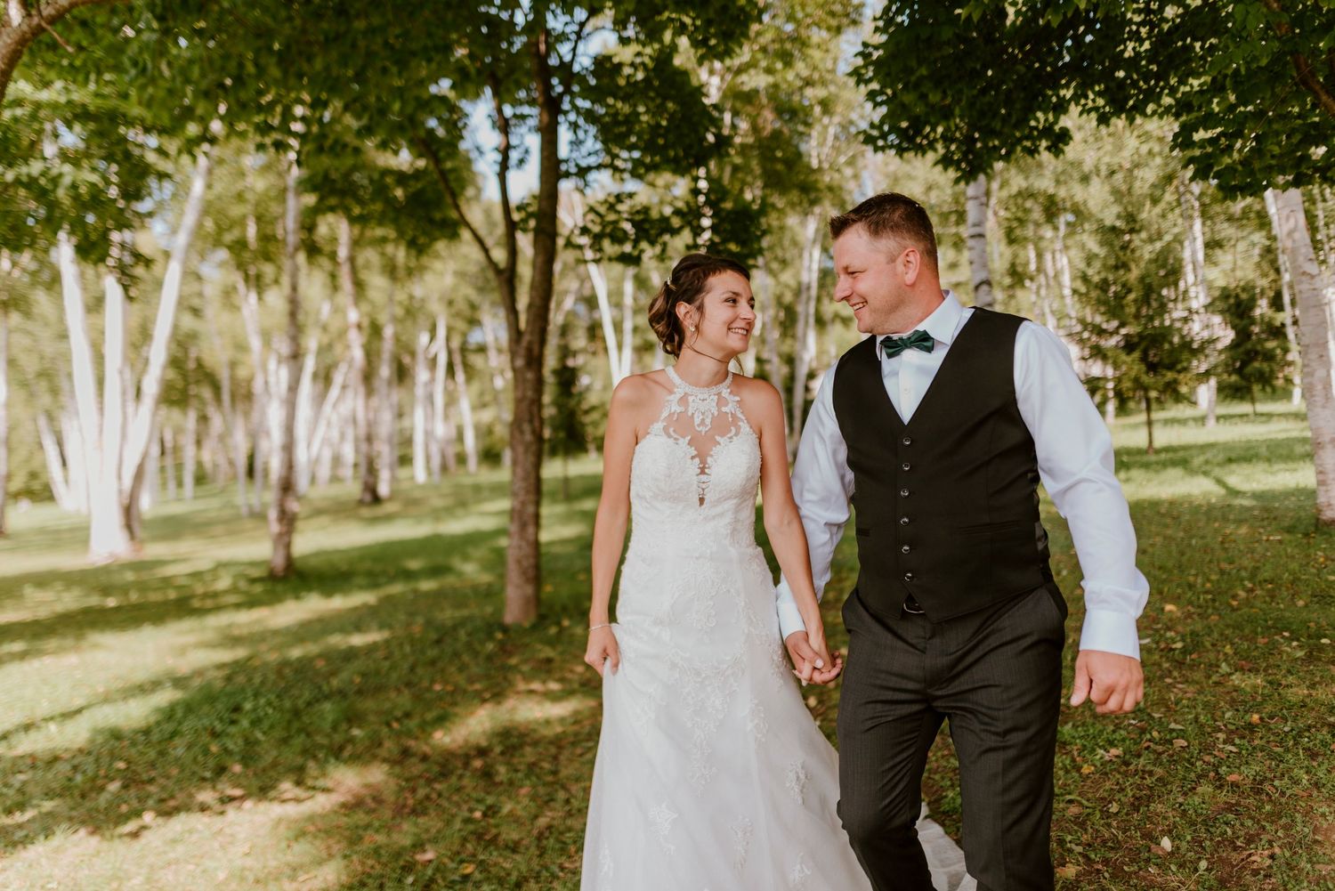 Casual wedding portrait of couple walking hand in hand through sunlit wooded area.