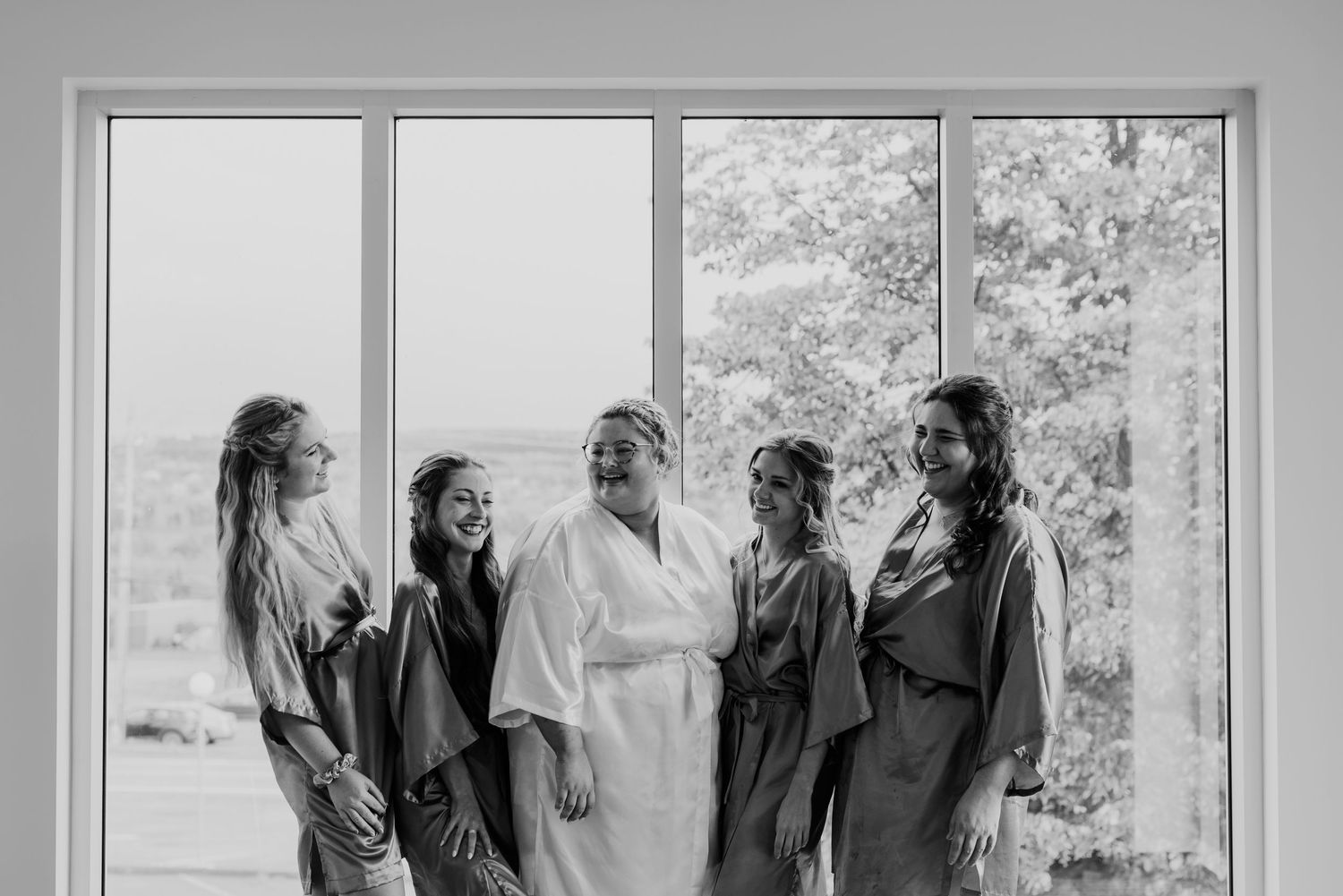 Group of wedding party members in robes standing by large windows during wedding preparations.