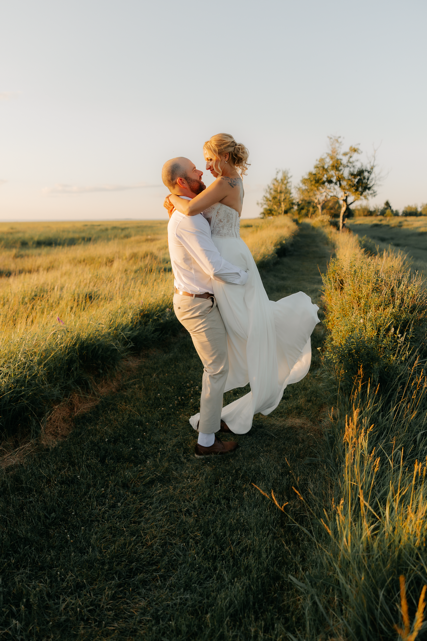 Groom lifts bride in white dress at golden sunset in a lush green field during outdoor wedding photo session.