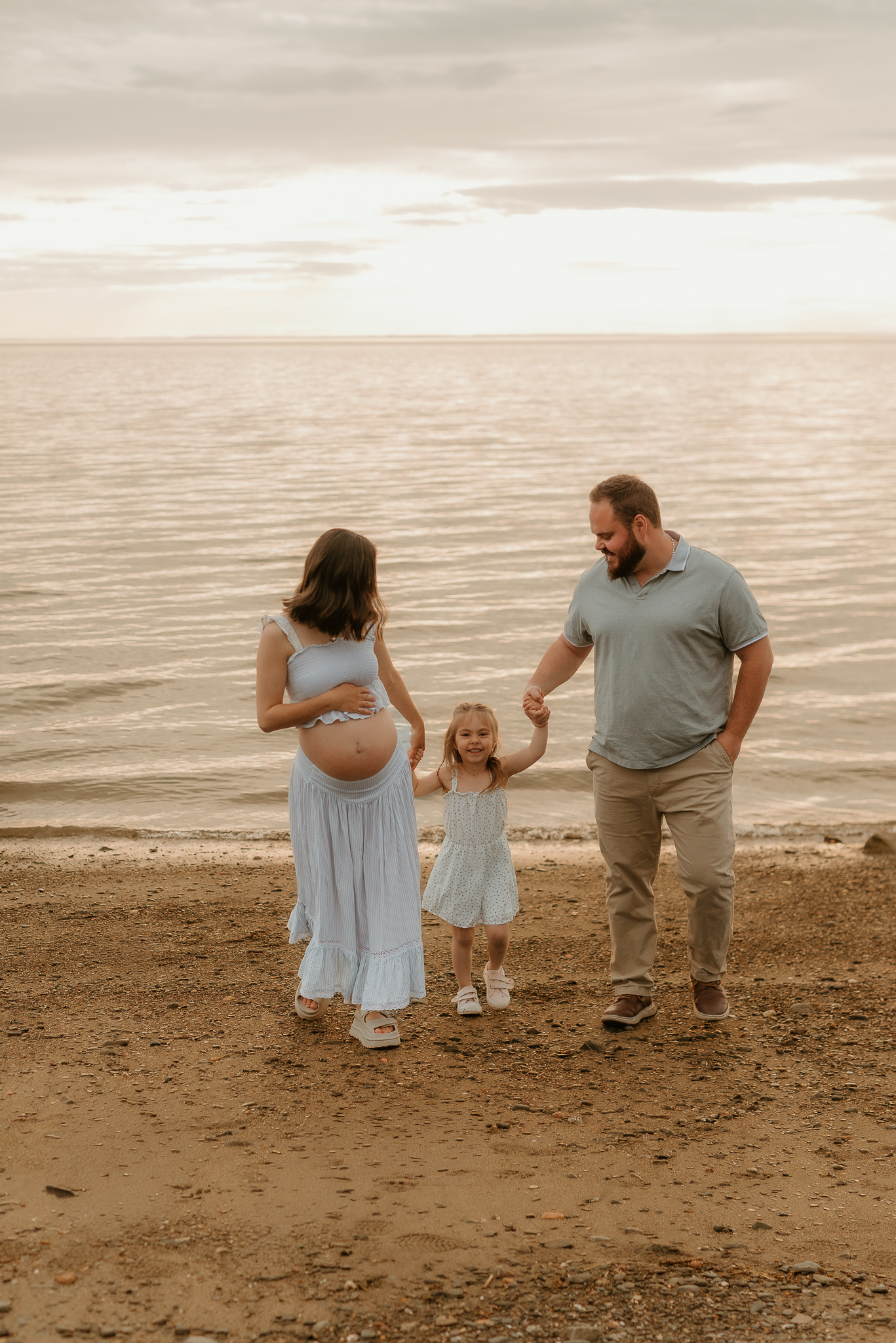 Pregnant woman in blue dress with husband and toddler daughter holding hands on sandy beach at sunset.