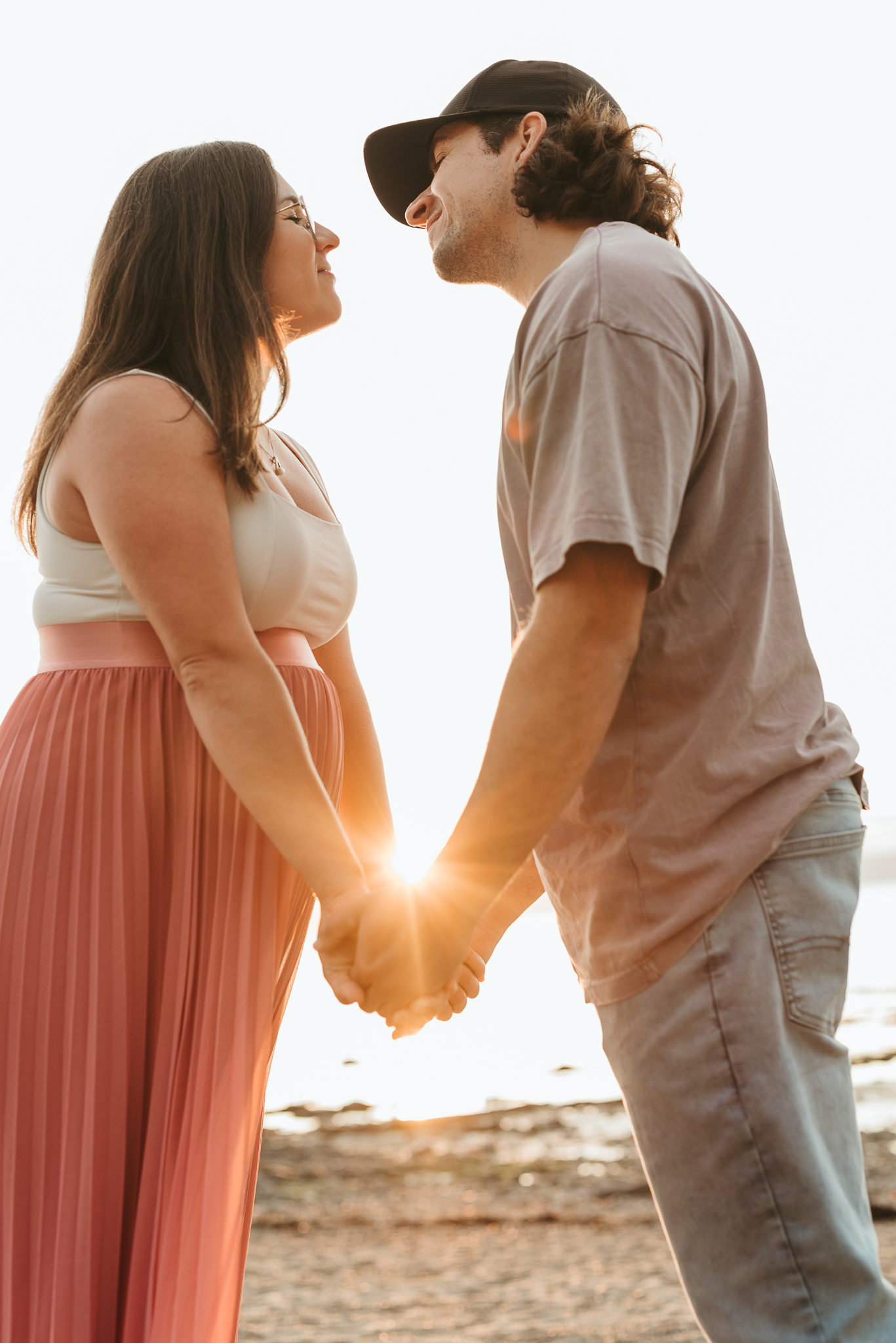 Couple holding hands at sunset on a beach, smiling at each other, woman in pink skirt and man in cap.