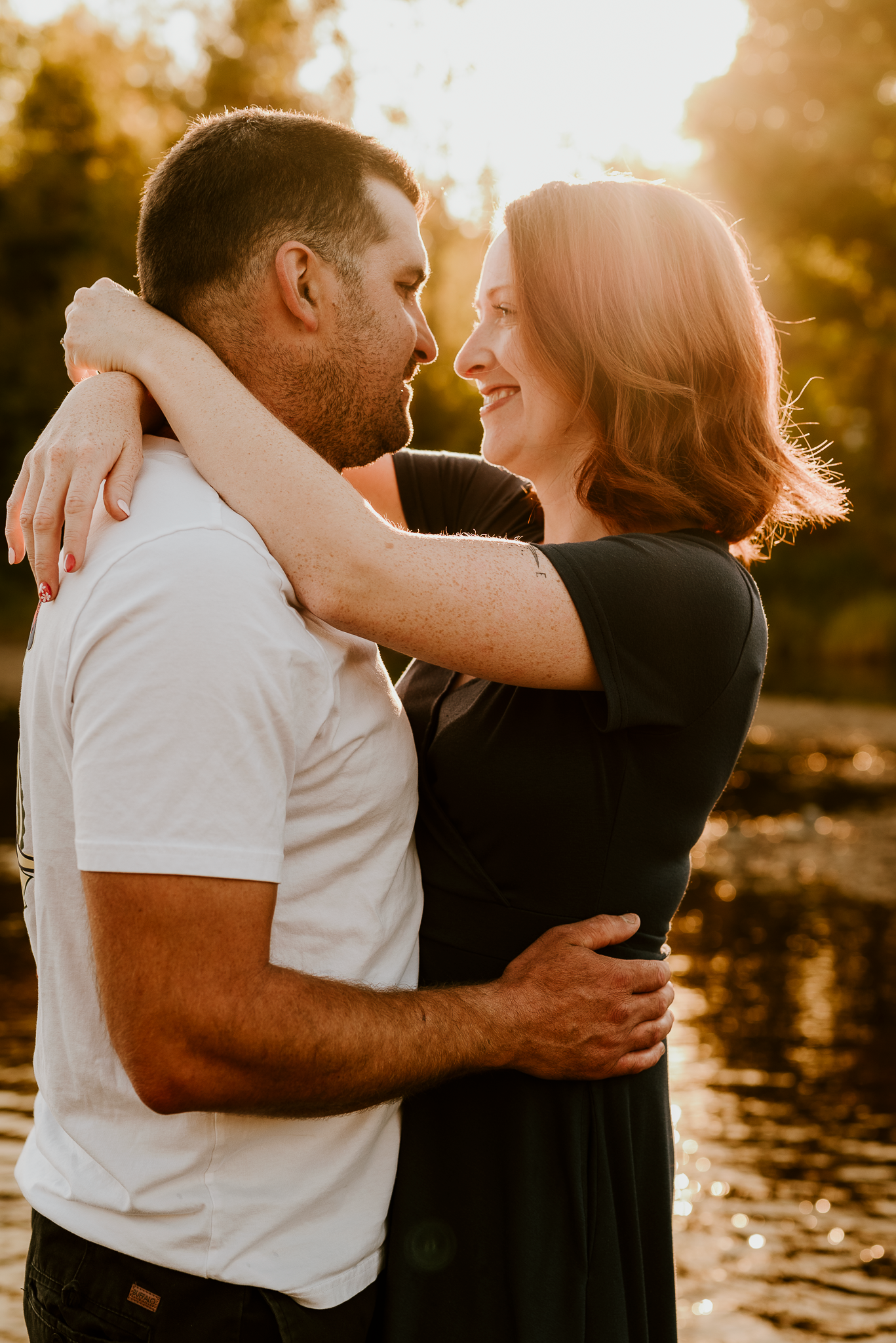 A couple embraces and smiles at each other during a golden hour outdoor photo session near a sunlit lake.