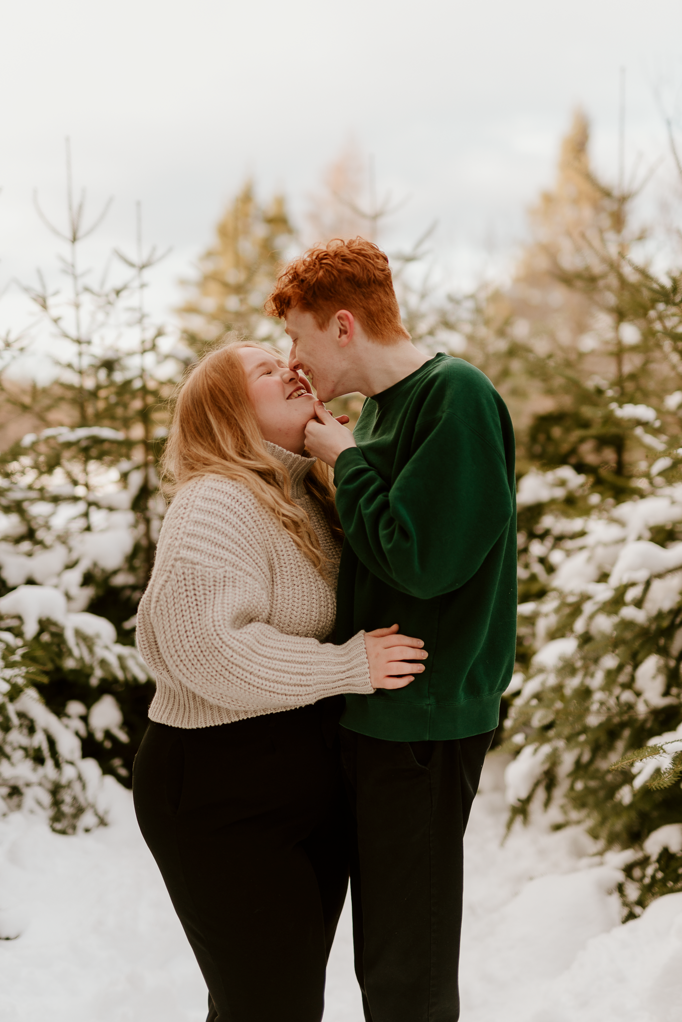 A couple embraces and smiles at each other among snow-covered evergreen trees in a cozy winter outdoor photo session.
