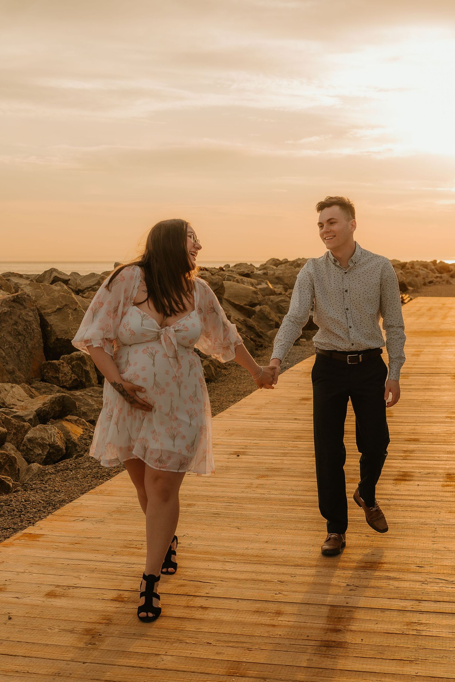 Couple holding hands walking on a wooden boardwalk by rocky shore at golden sunset.