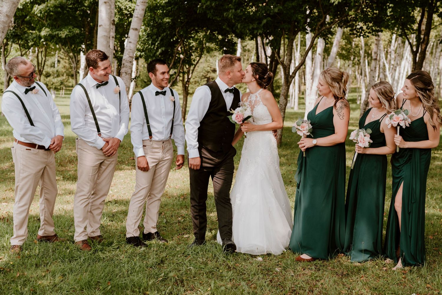 Wedding party lineup with groomsmen in suspenders and bridesmaids in emerald green dresses.