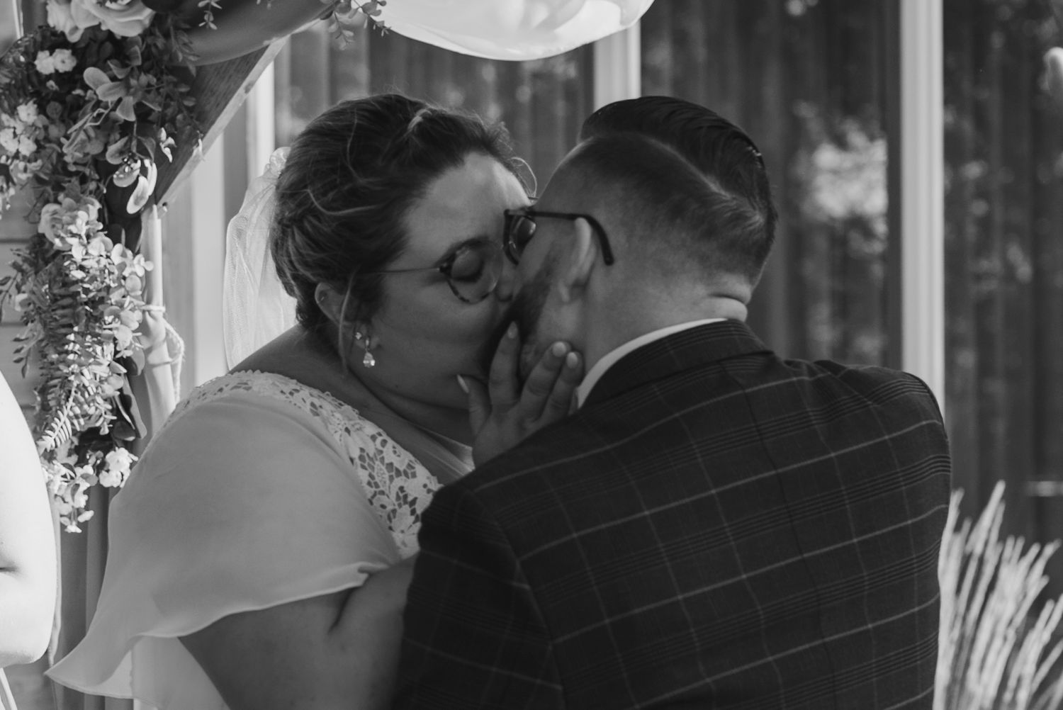 Intimate moment between newlyweds under floral arch in black and white.