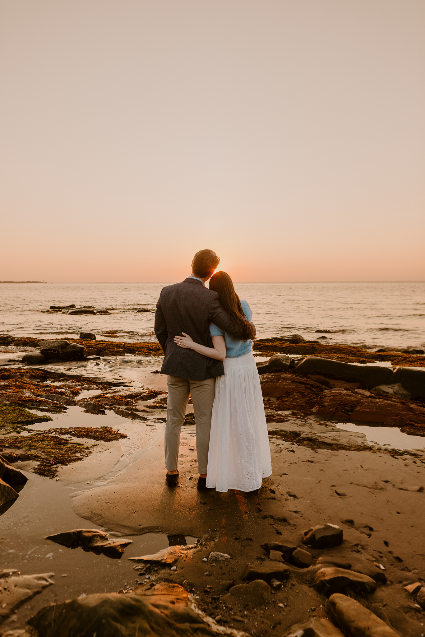 Couple embracing at rocky beach during golden sunset, woman in white dress and man in suit facing the ocean.