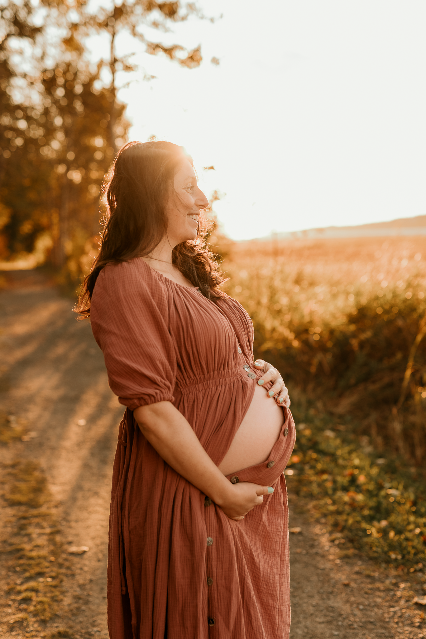 Smiling pregnant woman in rust dress holds baby bump on golden sunset dirt road surrounded by autumn fields.