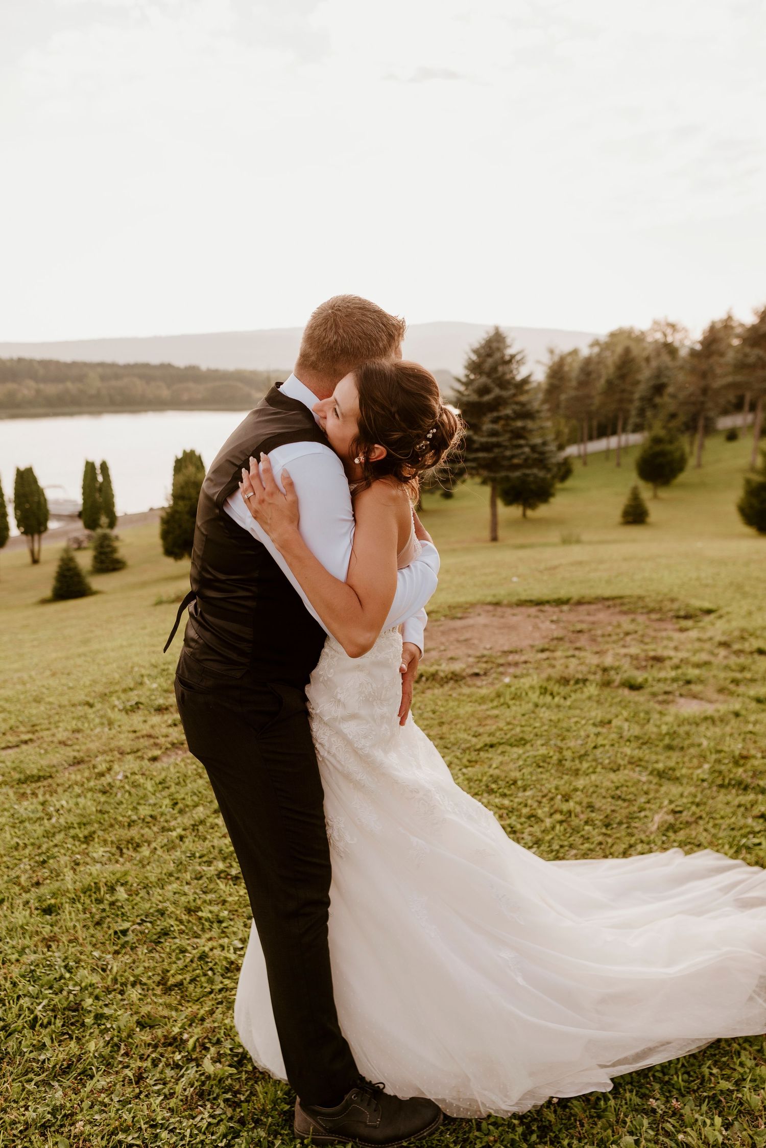 Intimate sunset portrait of newlyweds embracing on grassy hillside overlooking water.