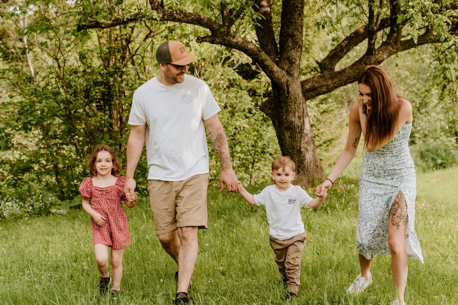 Family of four holding hands walking through lush green meadow with trees in background on sunny day.