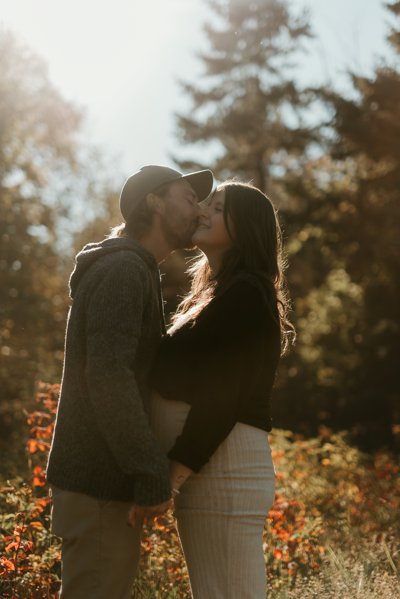 Couple sharing a romantic kiss outdoors in golden sunlight surrounded by autumn foliage.