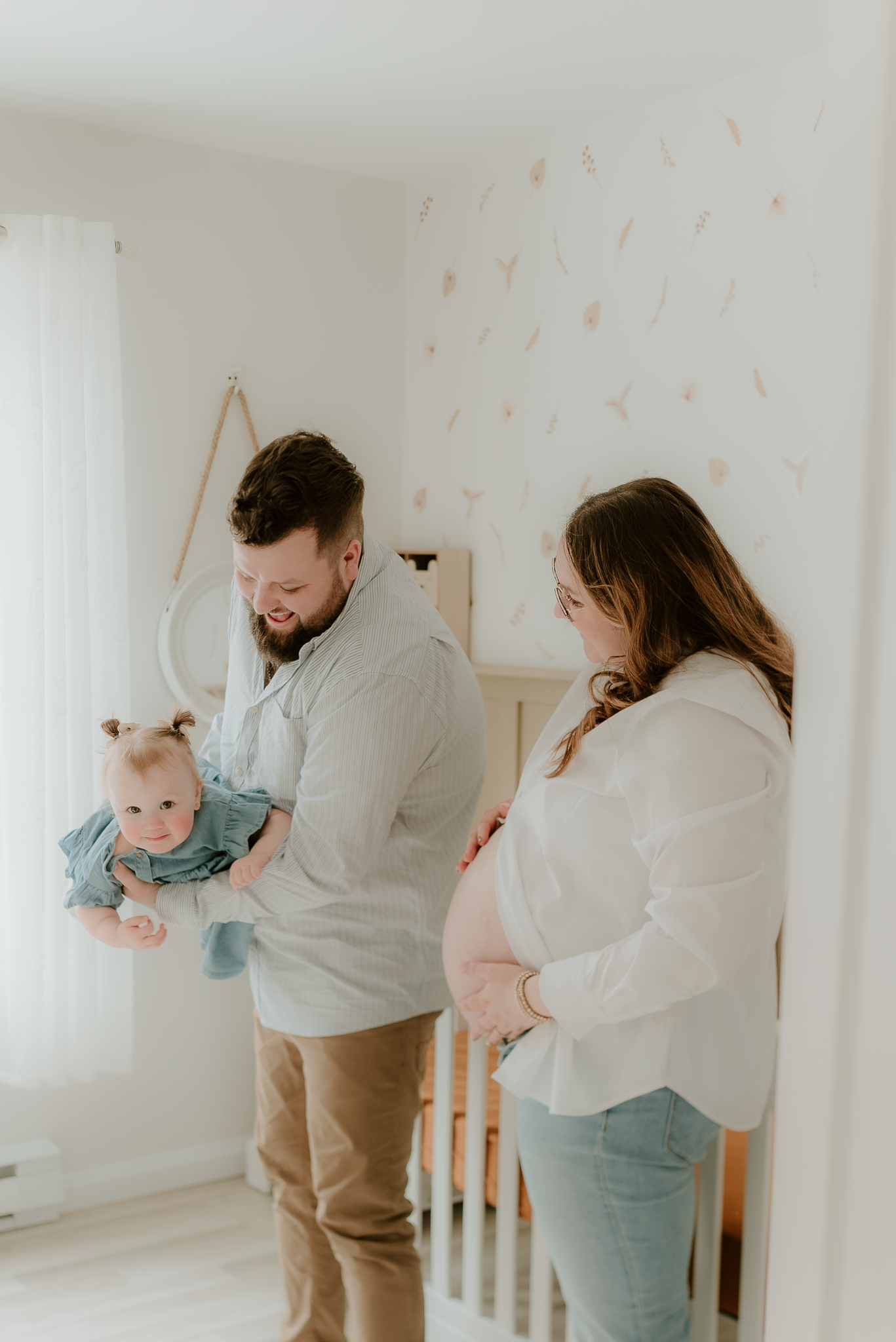 A couple stands in a nursery, man holds a baby girl while pregnant woman cradles her belly, soft white decor.