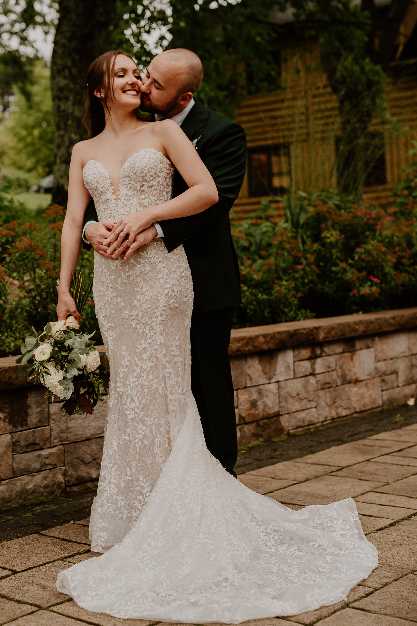 Bride in fitted lace gown holds bouquet while groom embraces her from behind.