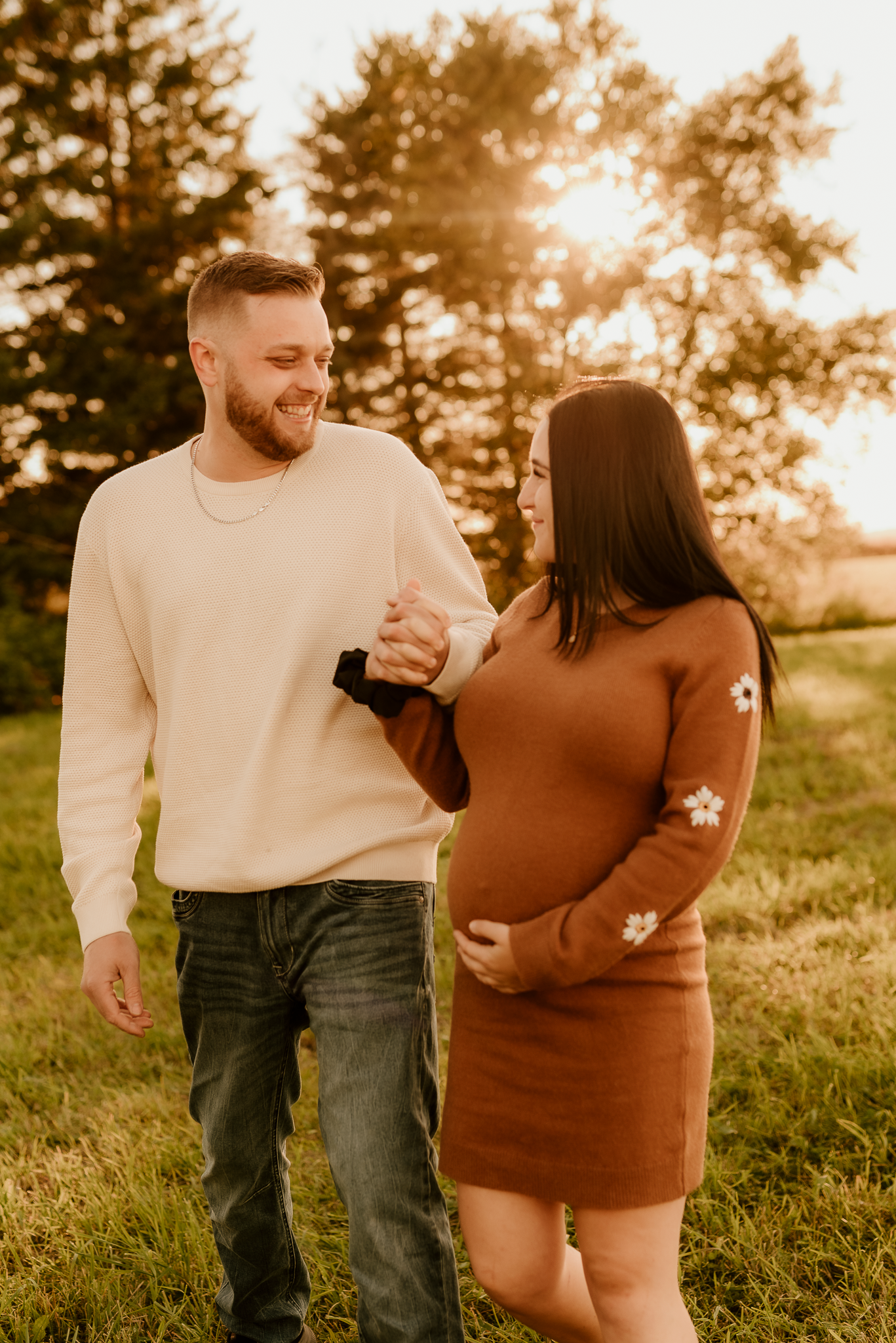 A couple shares a tender moment at sunset in a grassy field with pine trees in the background.
