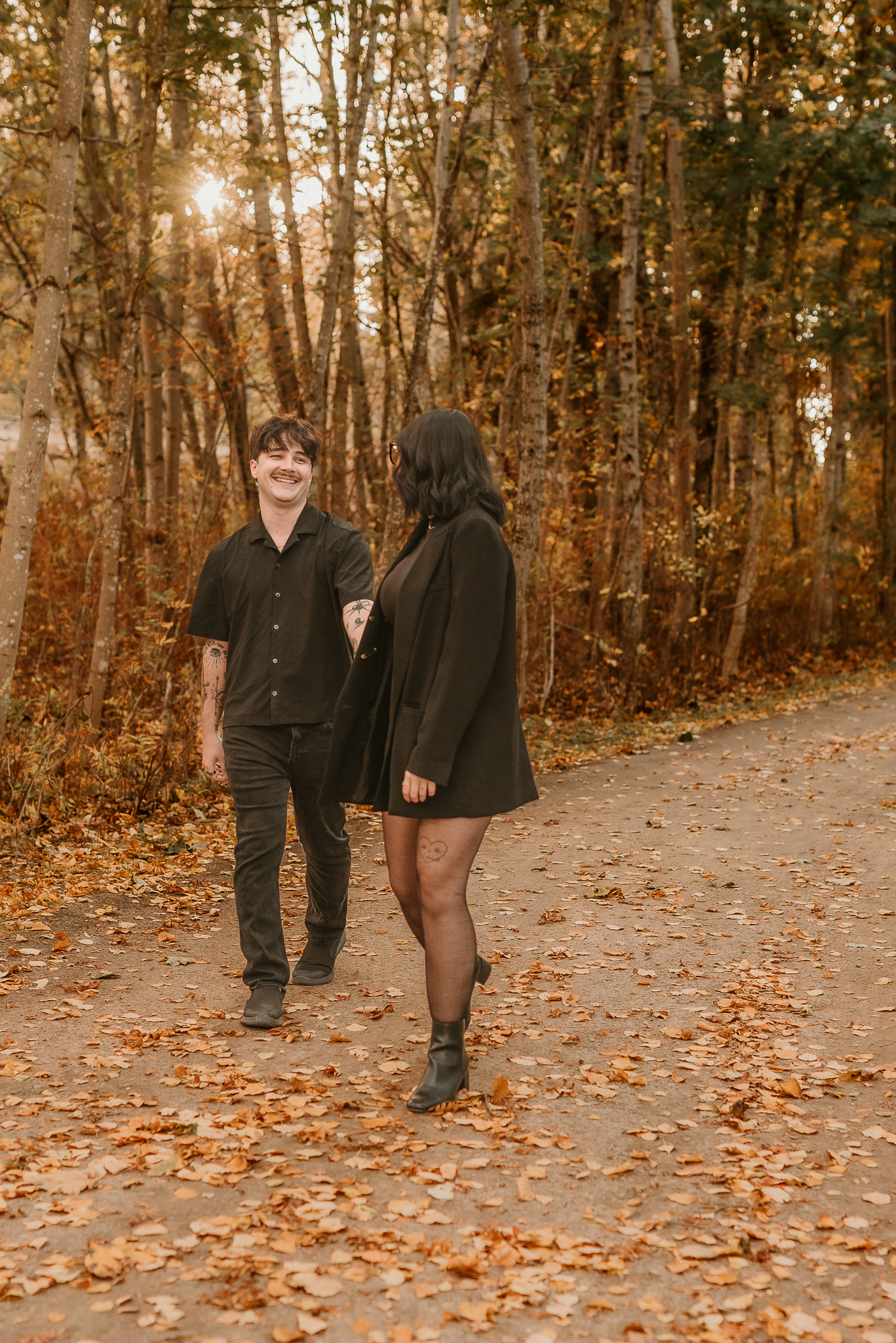Couple walking hand in hand on a leaf-covered path in an autumn forest, both dressed in black outfits.
