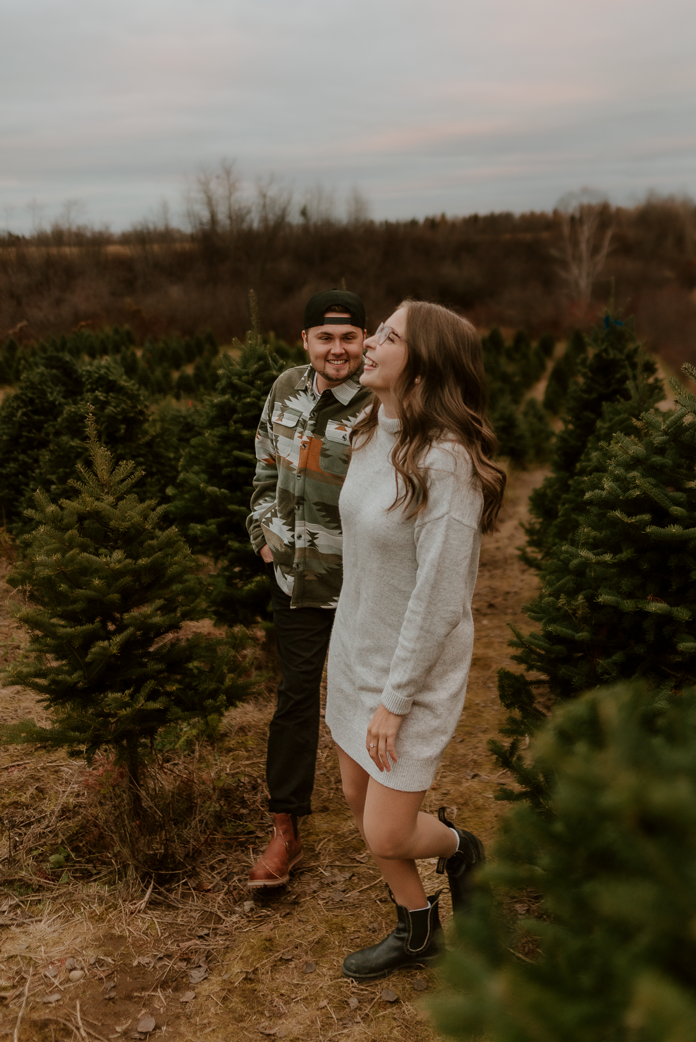 Couple laughing together at a Christmas tree farm, woman in white sweater dress and man in plaid shirt among evergreen trees.
