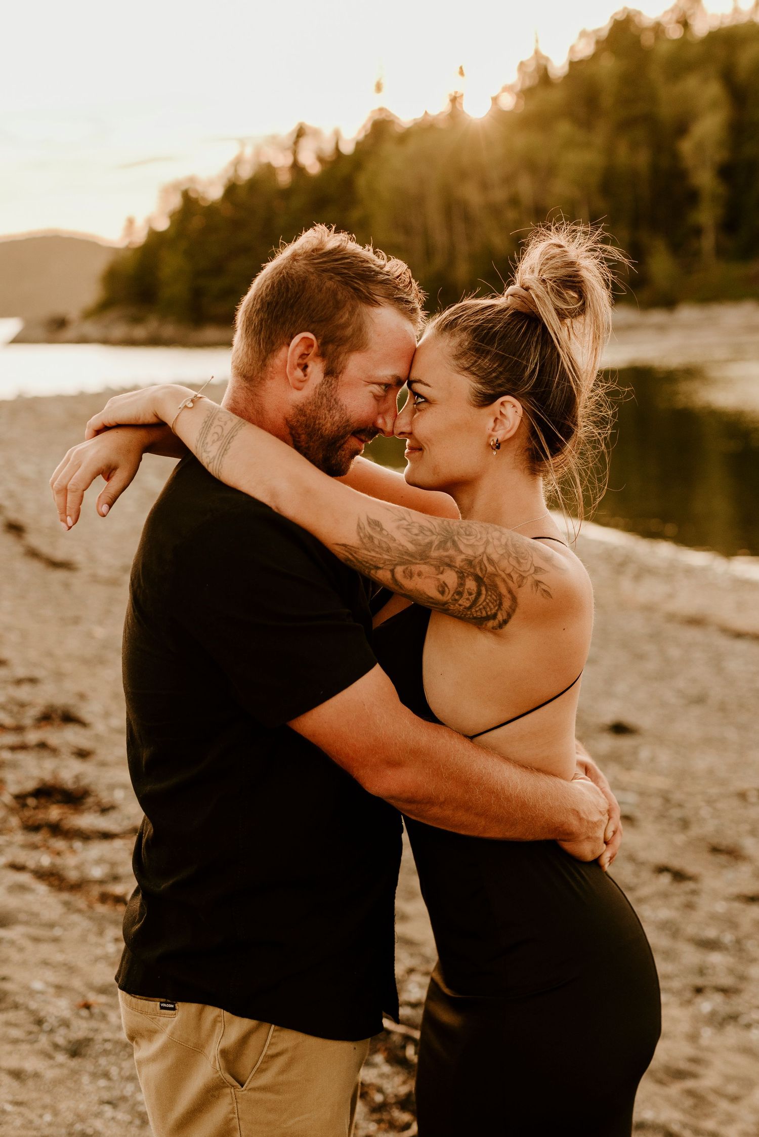 A couple with tattoos embraces tenderly on a beach at golden hour with a forested hillside in the background.