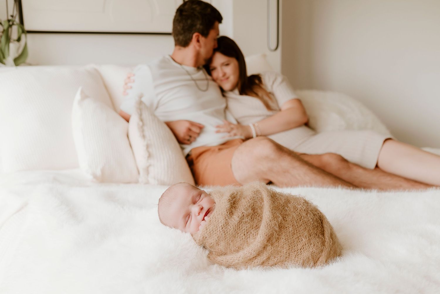 Couple relaxing on white bed gazing at their newborn baby wrapped in a tan swaddle during an in-home newborn session.