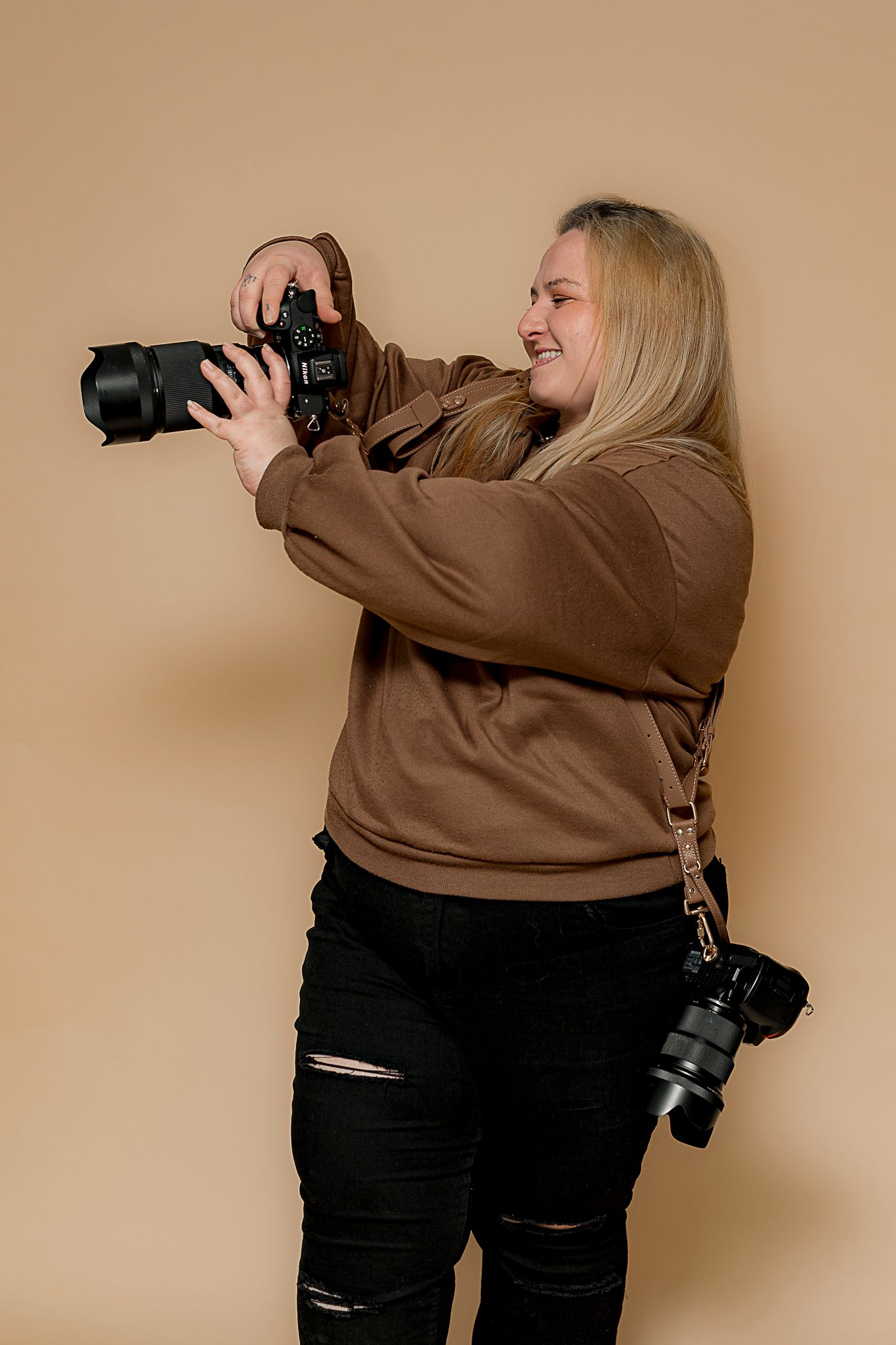 Blonde female photographer in brown hoodie and black ripped jeans holding DSLR camera, smiling against beige background.