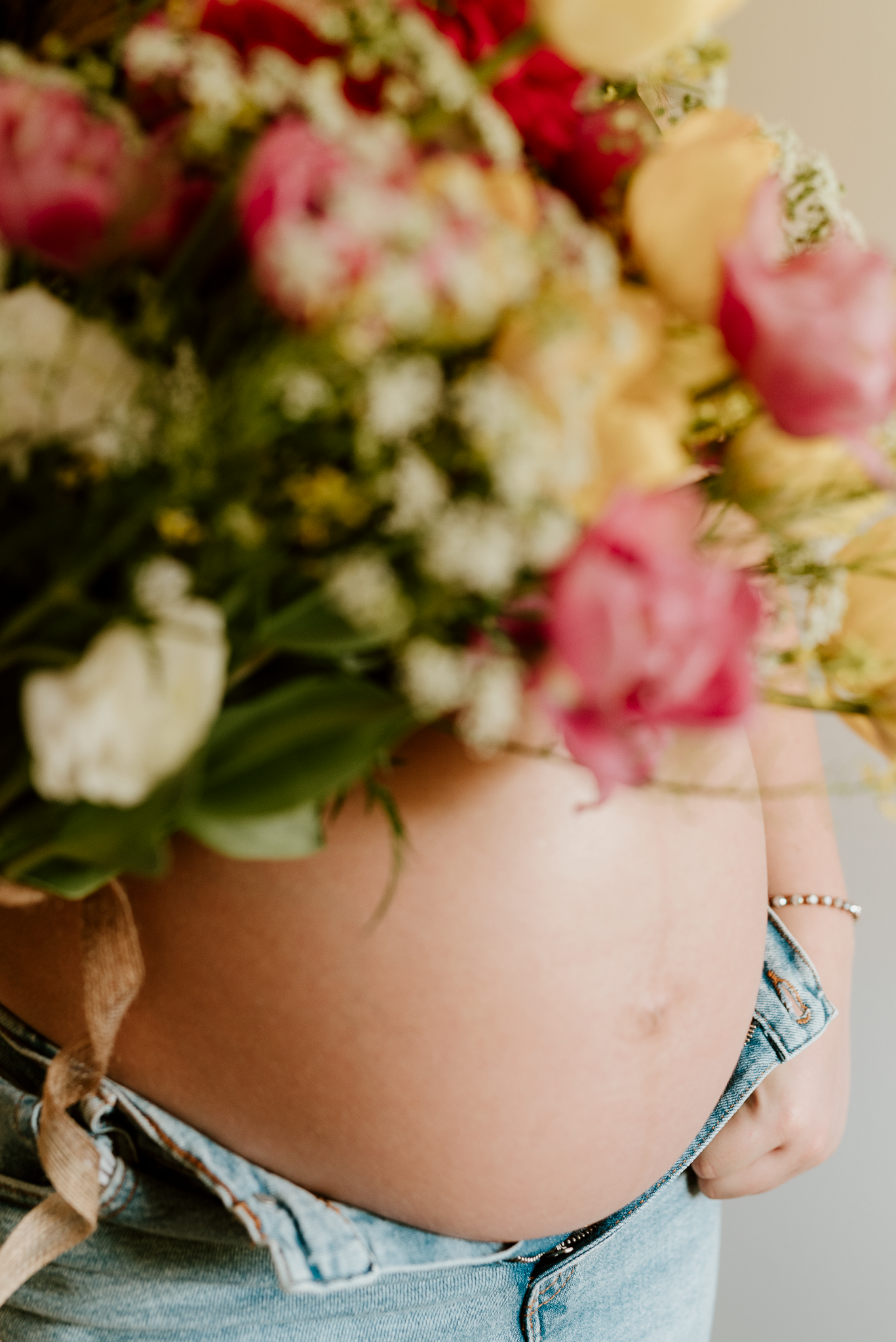 Pregnant woman's bare baby bump with unbuttoned jeans, holding a colorful bouquet of pink, white, and yellow flowers.