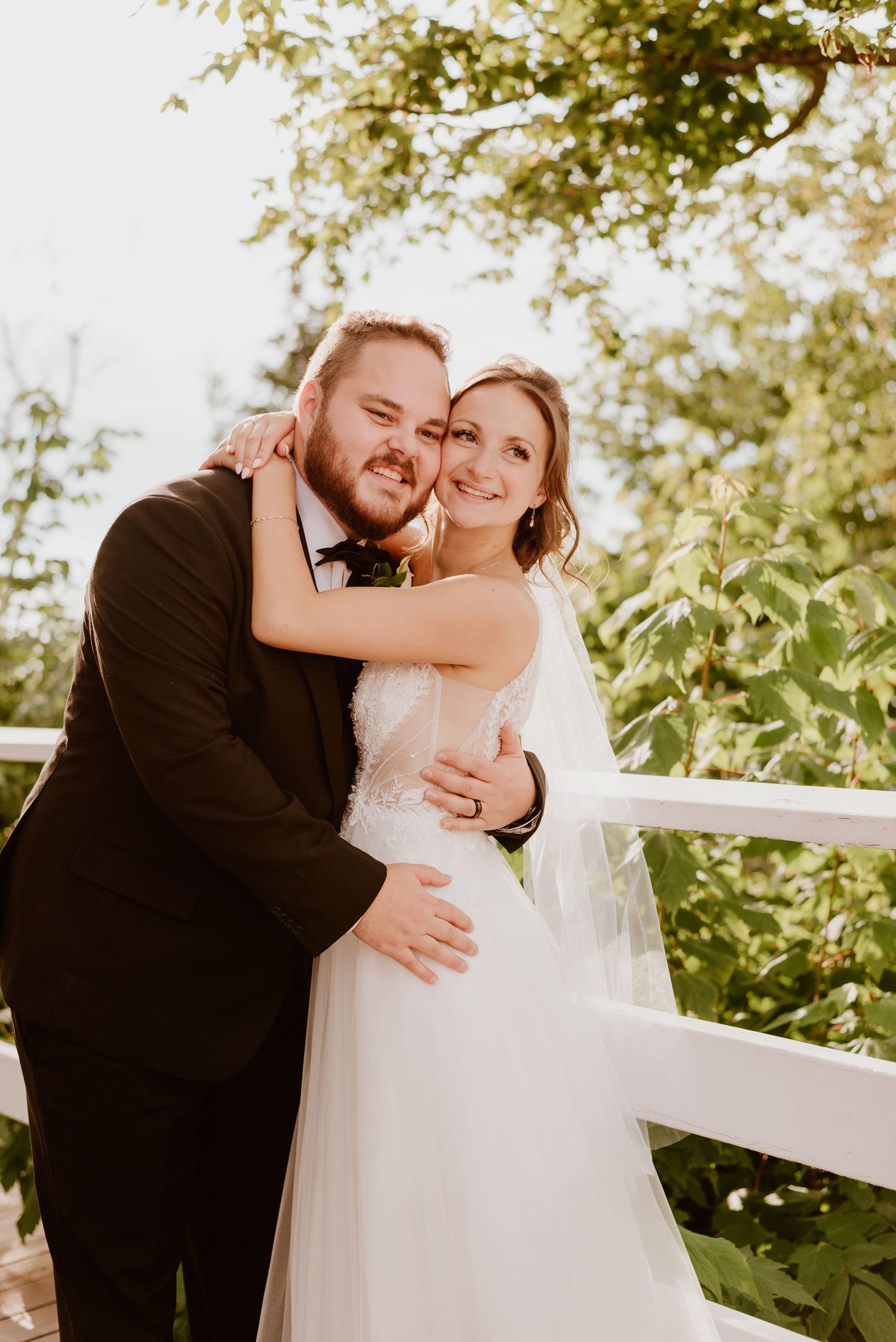 Bride and groom embrace on a white railed deck surrounded by lush green trees in warm golden sunlight.