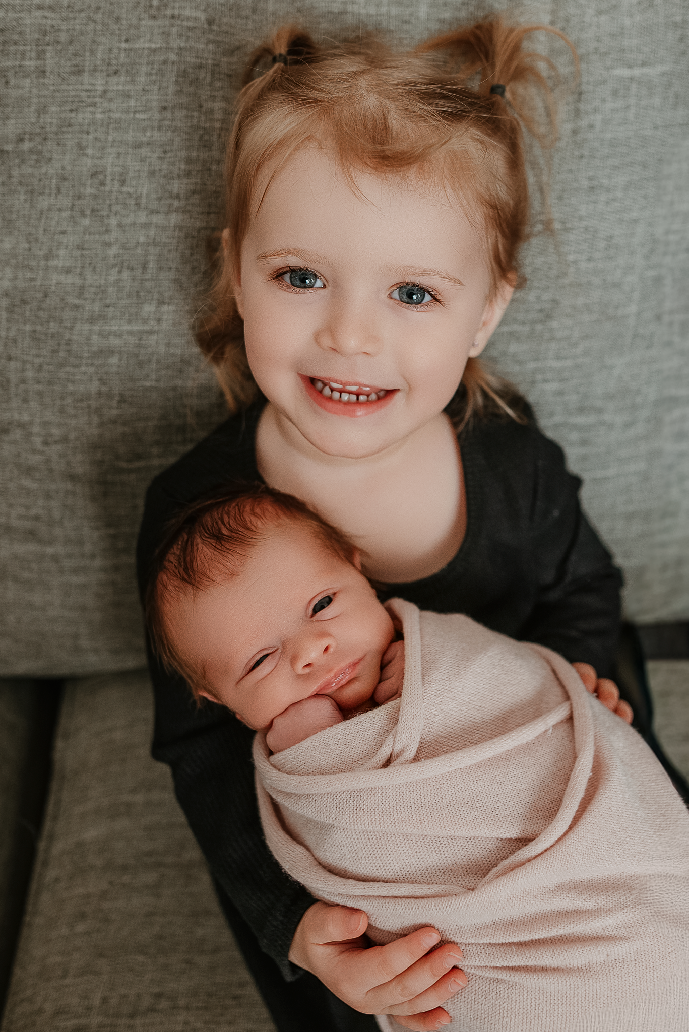 Young blonde toddler girl smiling and holding a swaddled newborn baby sibling on a gray couch.
