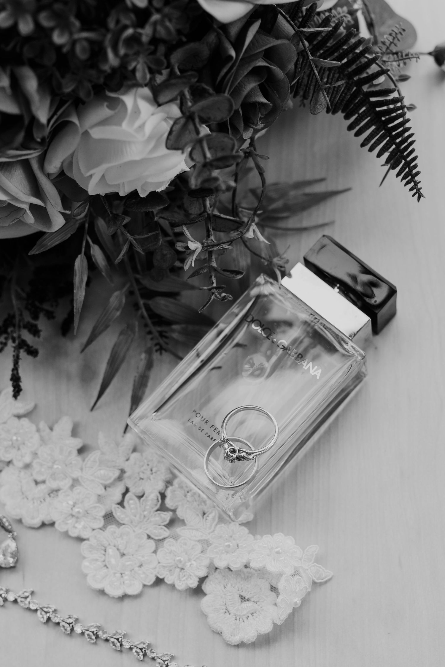Wedding rings and perfume bottle arranged with floral decor and lace in black and white.