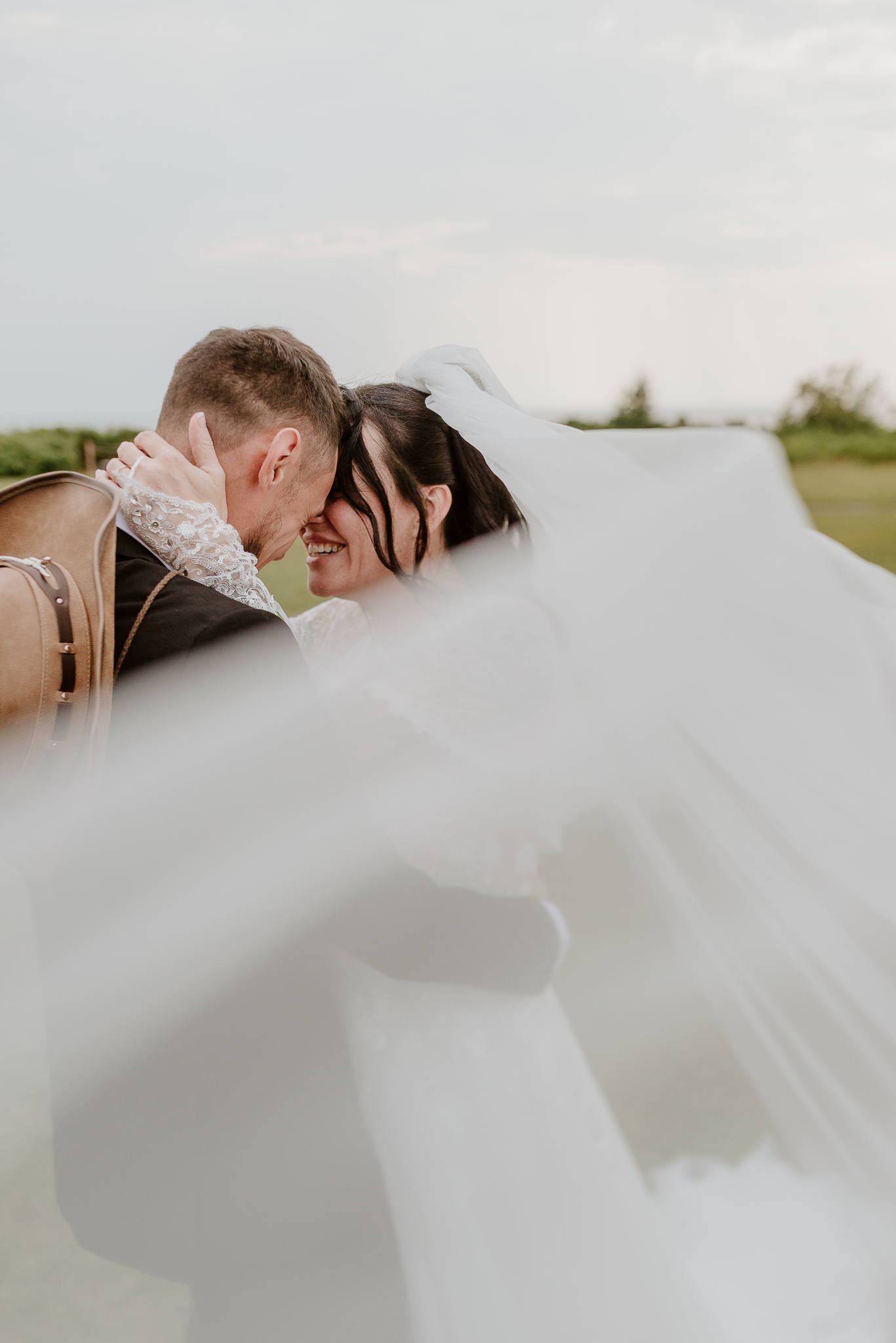 A bride and groom share a romantic moment under a flowing white veil outdoors on their wedding day.