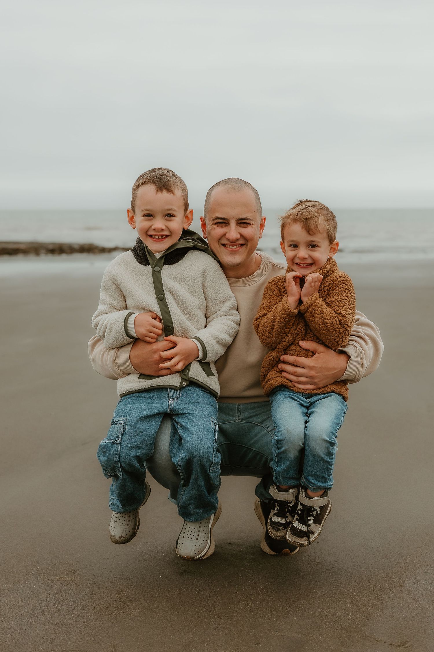A parent crouches on a sandy beach with two young boys, all smiling, wearing casual sweaters and jeans on an overcast day.