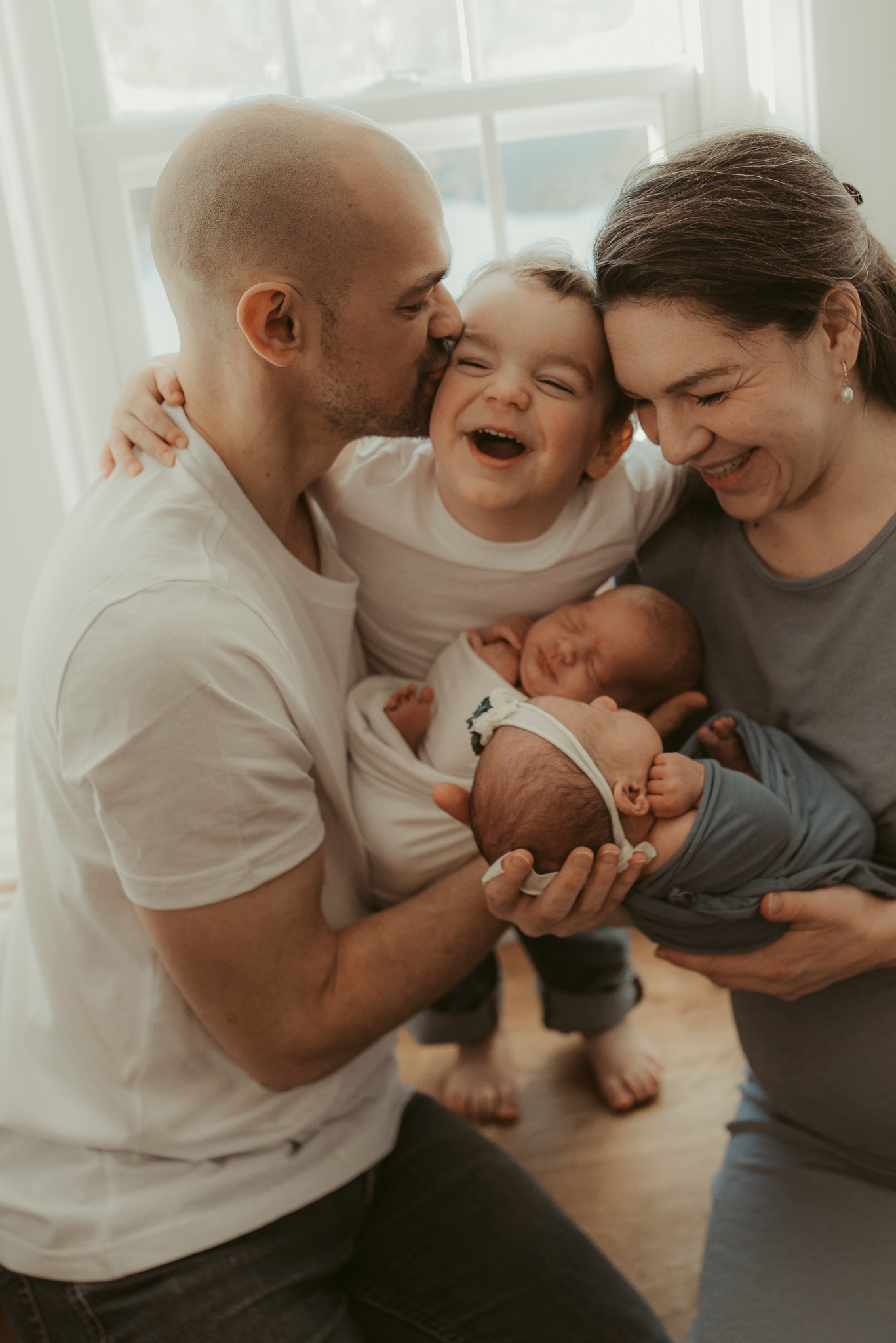 Happy family with parents smiling and kissing toddler while holding newborn baby near bright window.