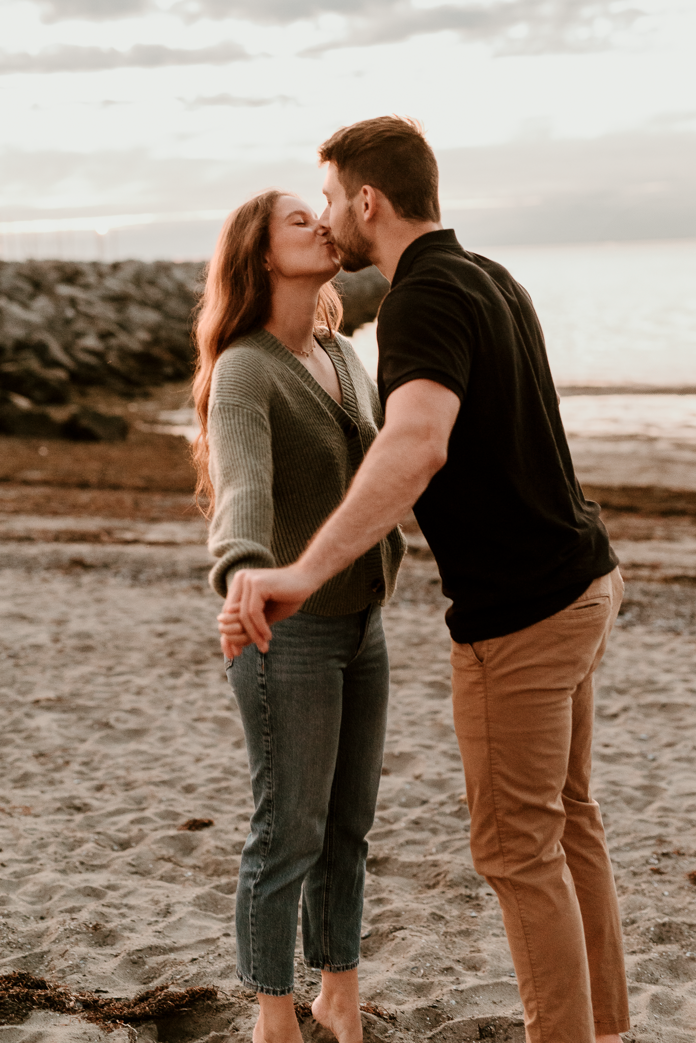 Couple sharing a romantic kiss on a sandy beach at sunset, holding hands near rocky shoreline.
