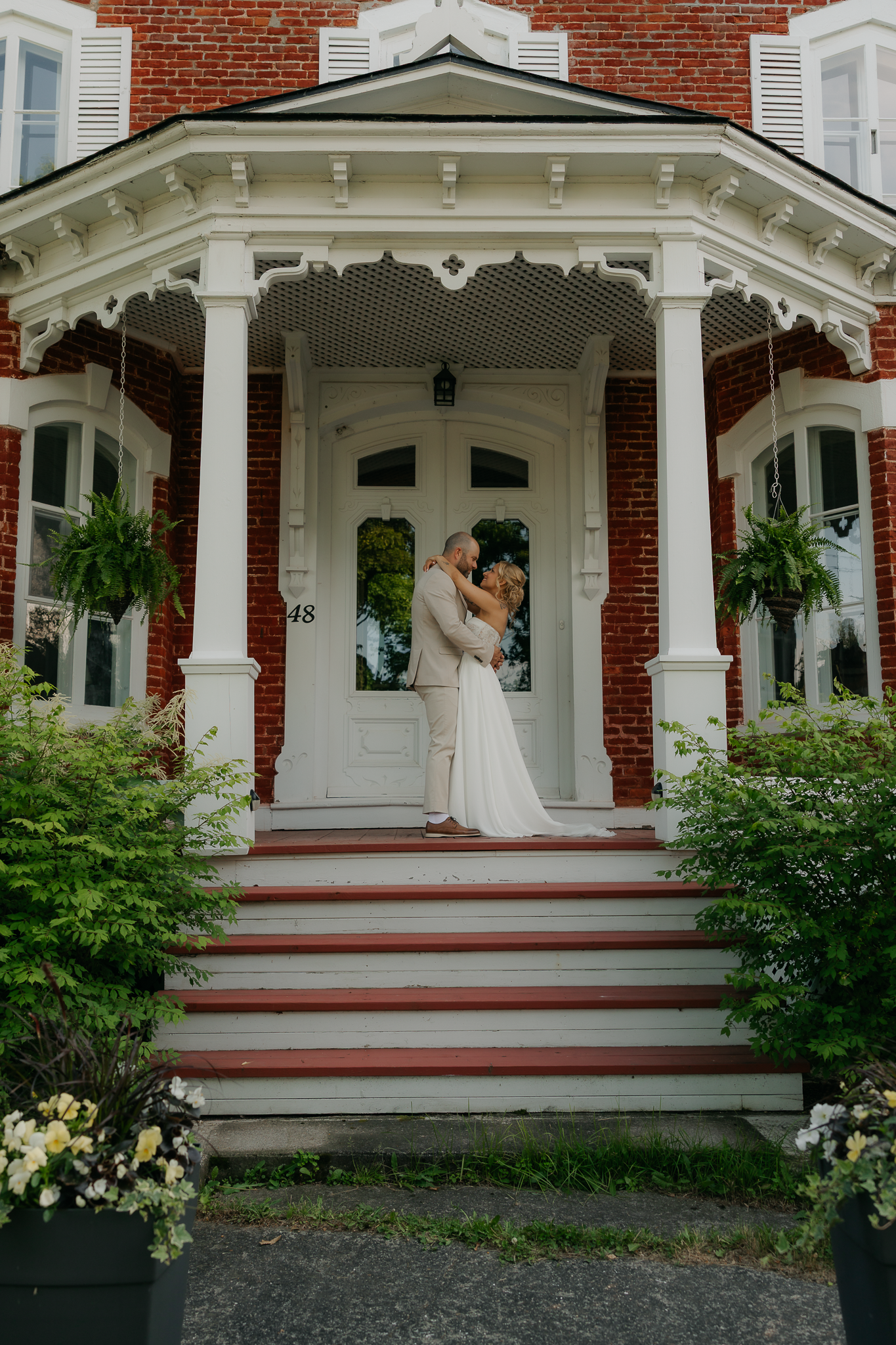 A couple shares a romantic kiss on the grand porch steps of a charming red brick Victorian home on their wedding day.